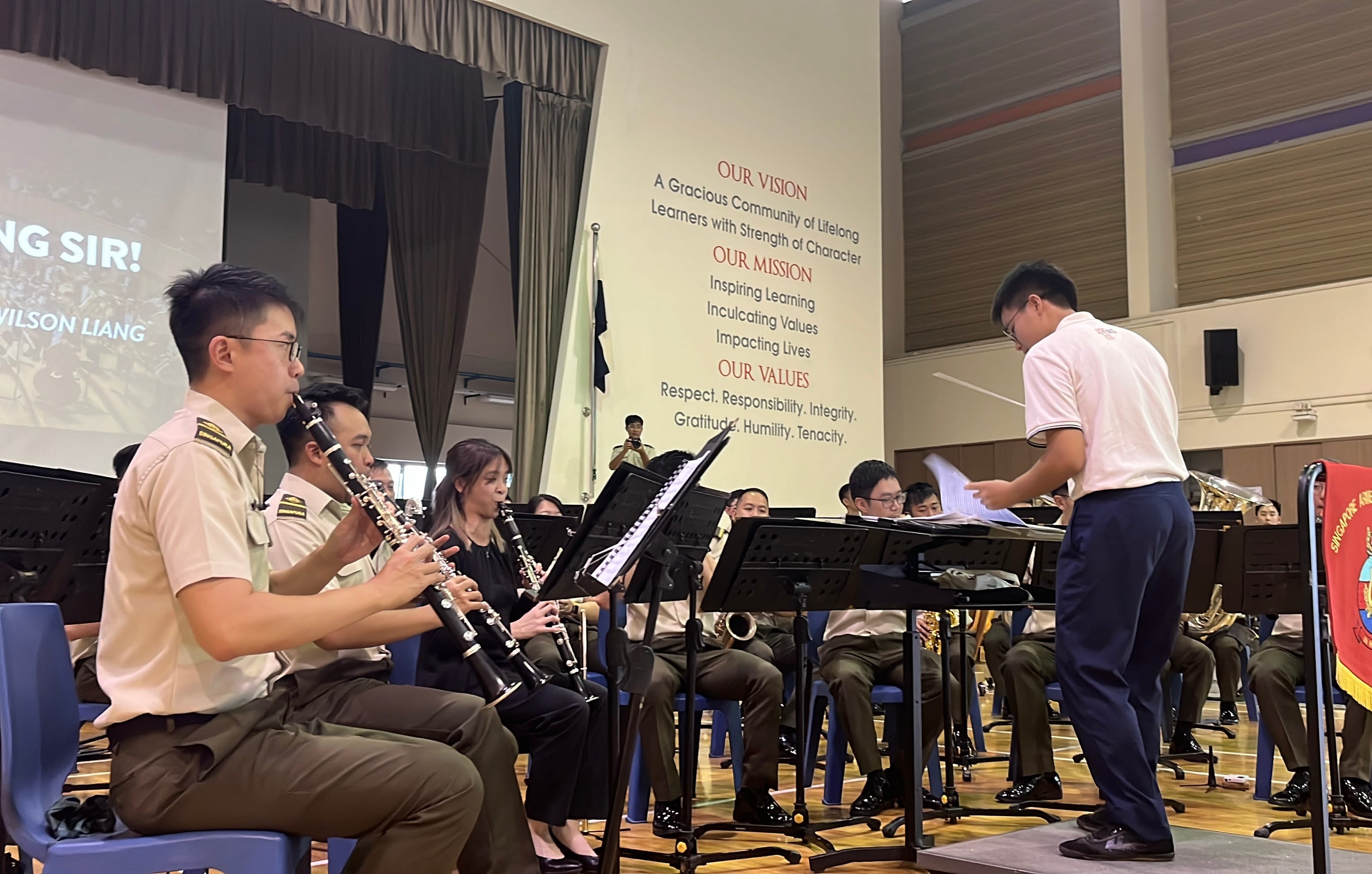 A uniformed band plays clarinets in a school hall while a conductor stands before music stands.