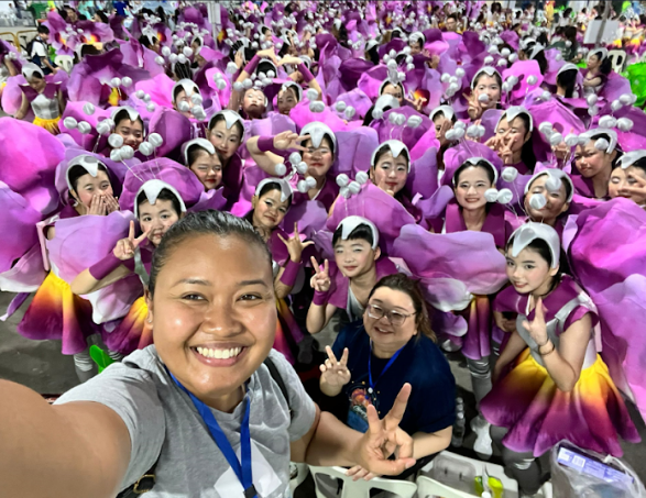 Selfie with a large group of performers wearing purple flower costumes and white petal headpieces, posing with peace signs.