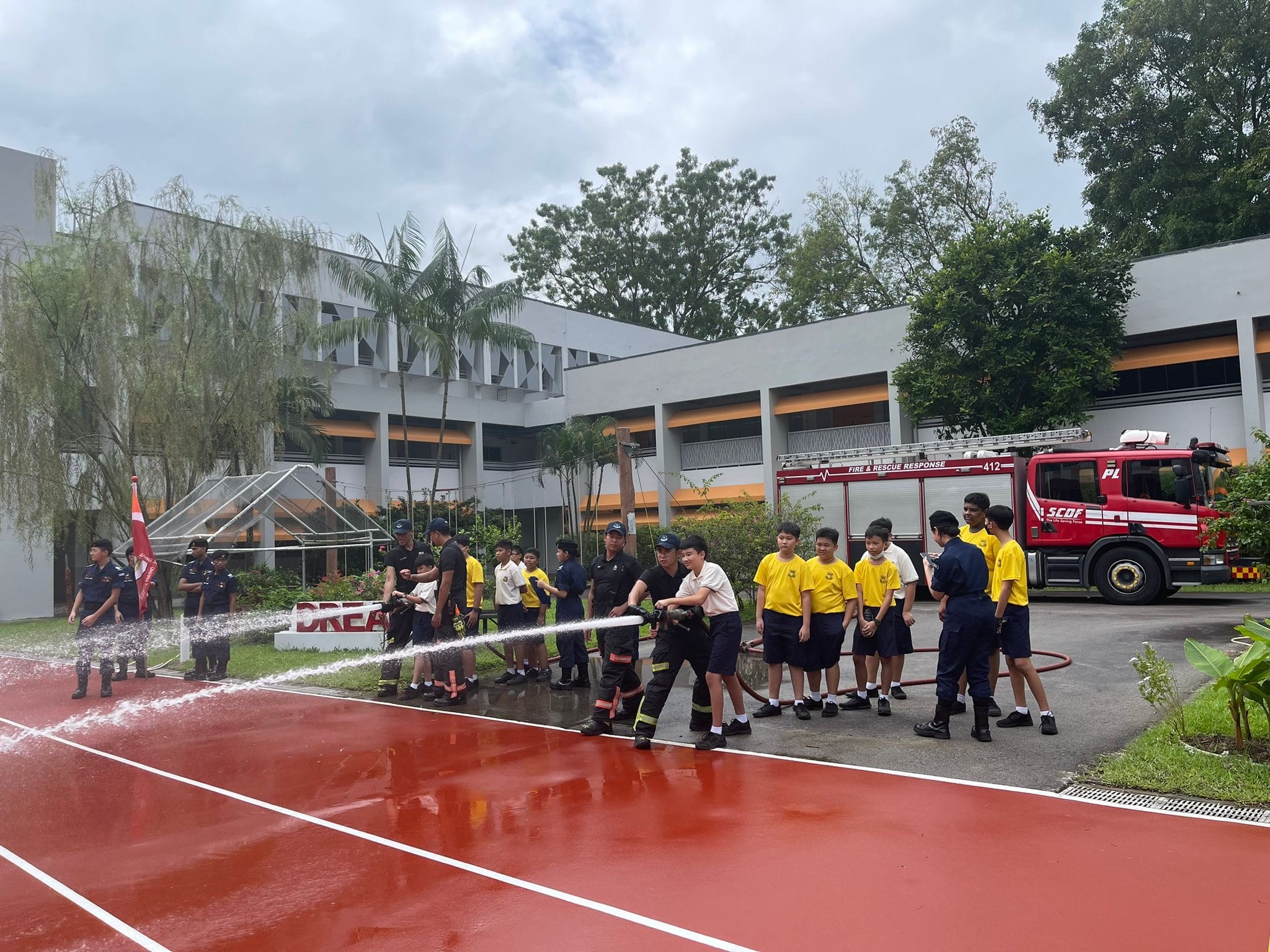 Students in yellow uniforms practice using a fire hose with SCDF firefighters on a school track, with a red fire truck nearby.