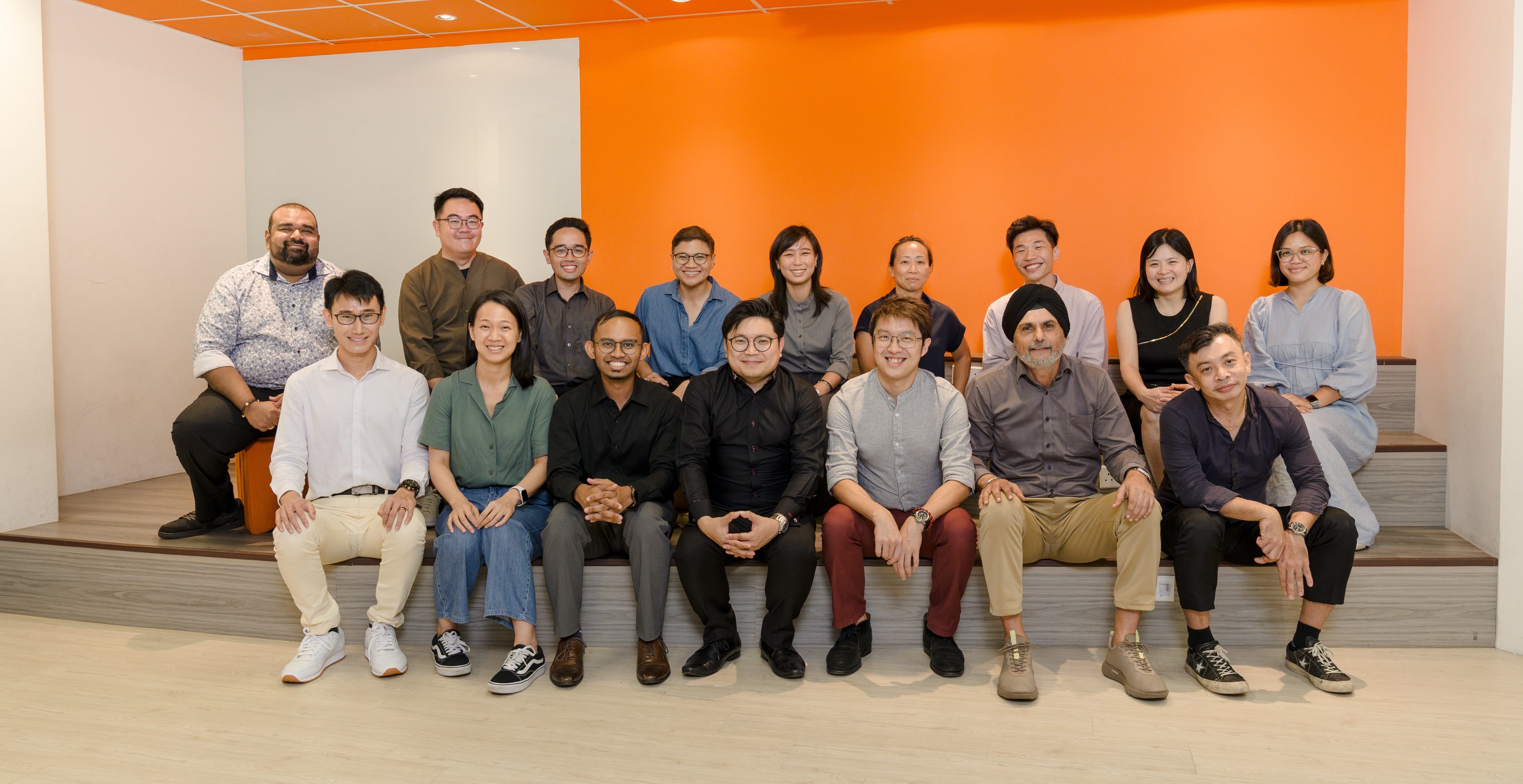 A group of about 16 people posing together on tiered steps in front of an orange wall indoors.