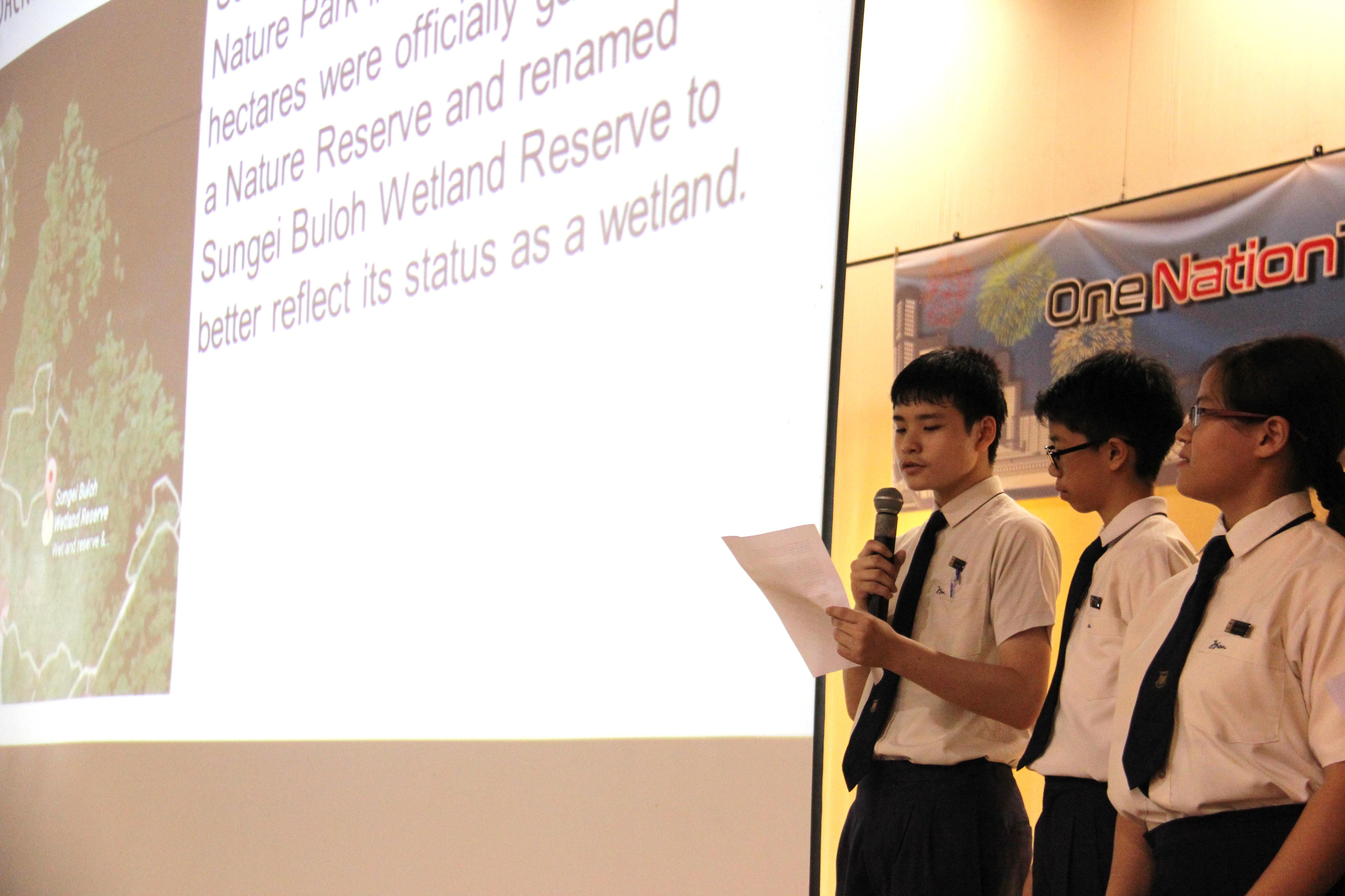 Three uniformed students present at a podium, one holding a microphone and paper before a Sungei Buloh Wetland Reserve slideshow.
