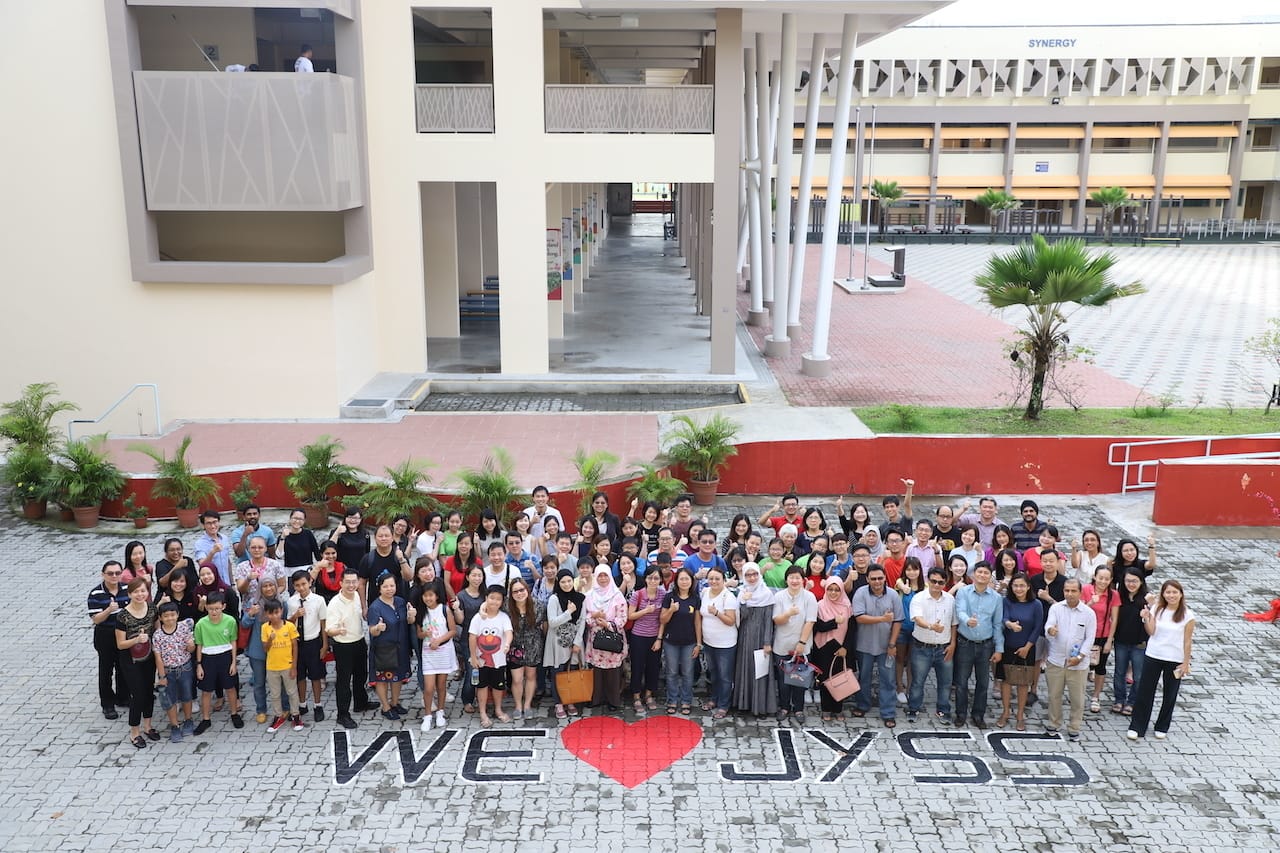 Large group of people posing outdoors at a school courtyard in front of a "WE LOVE JYSS" ground mural.