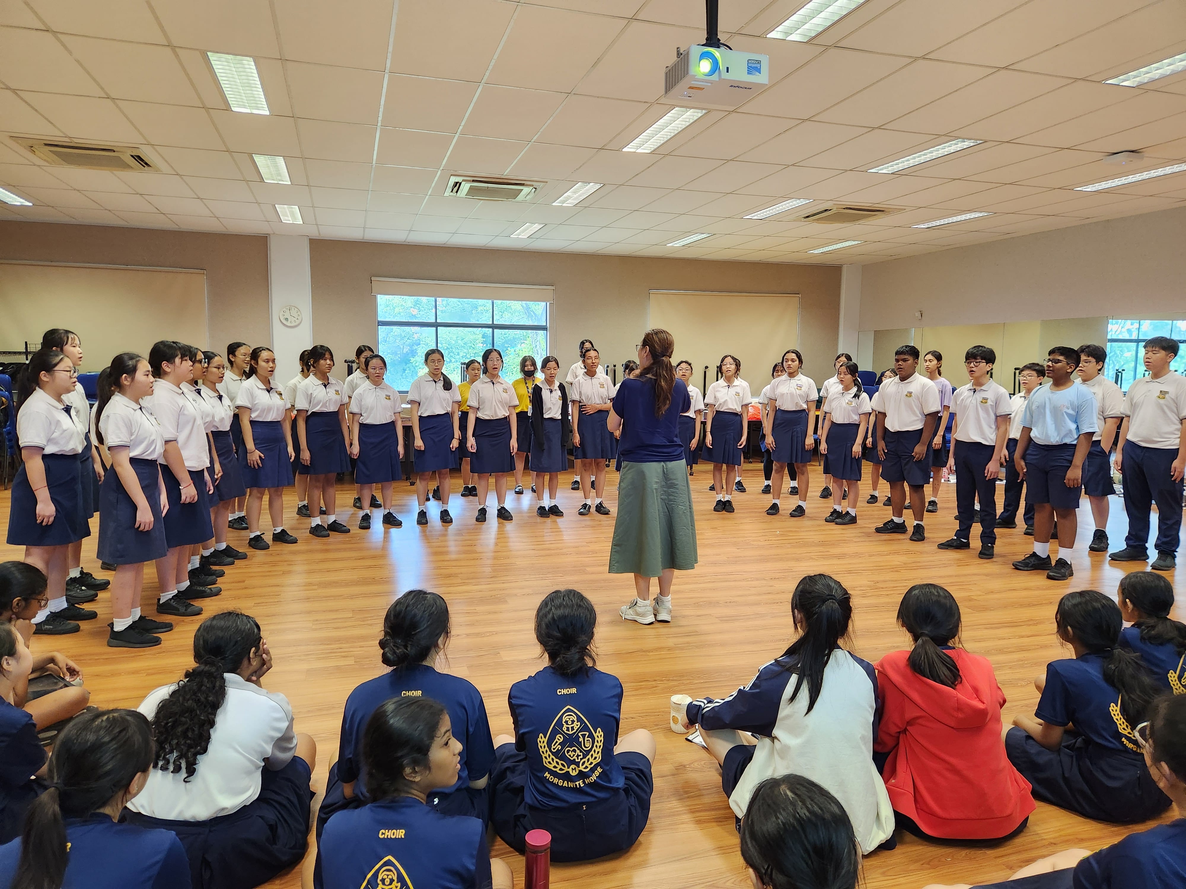 A conductor leads a large school choir rehearsal indoors, with students in uniform standing and others seated on the floor watching.