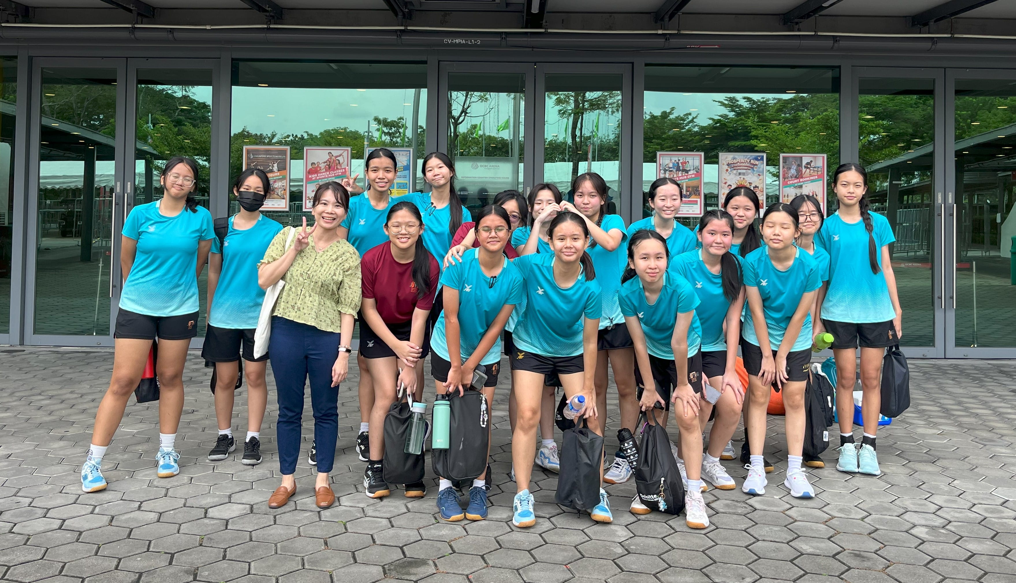 A group of teenage girls in matching teal sports uniforms pose with two adults outside a glass building.