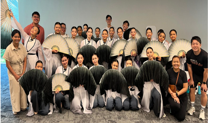 A large group of performers in white and gray costumes holding large decorative fans pose for a photo.