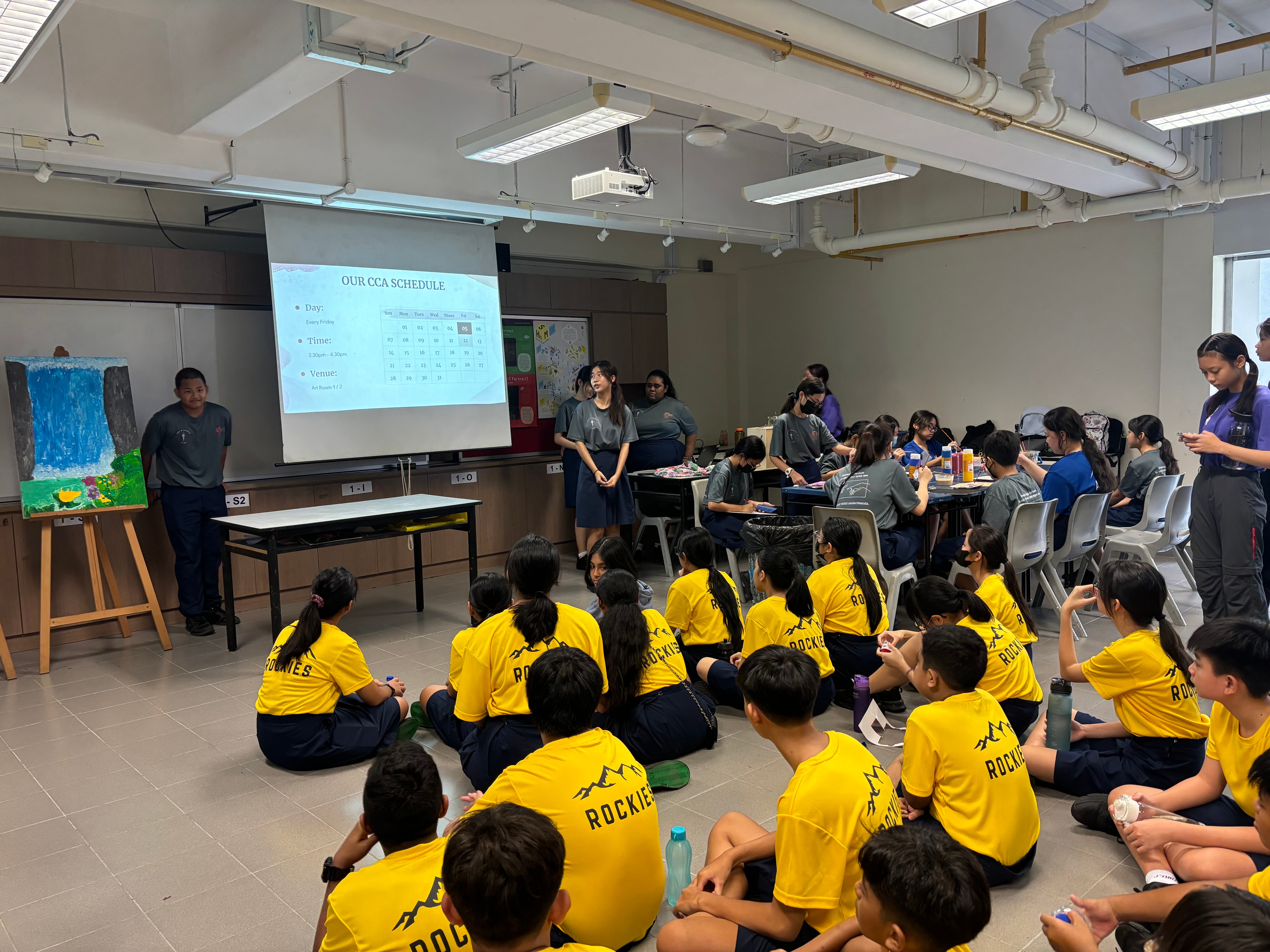 Children in yellow shirts sit on floor watching a presentation slide in a classroom while others sit at tables.