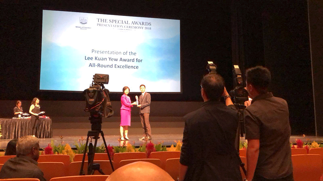 Two people exchange an award on stage at the 2018 Special Awards Ceremony, filmed by cameramen in an auditorium.