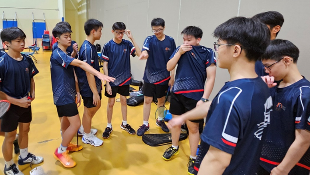 A group of young male badminton players in navy uniforms huddle together on an indoor court.