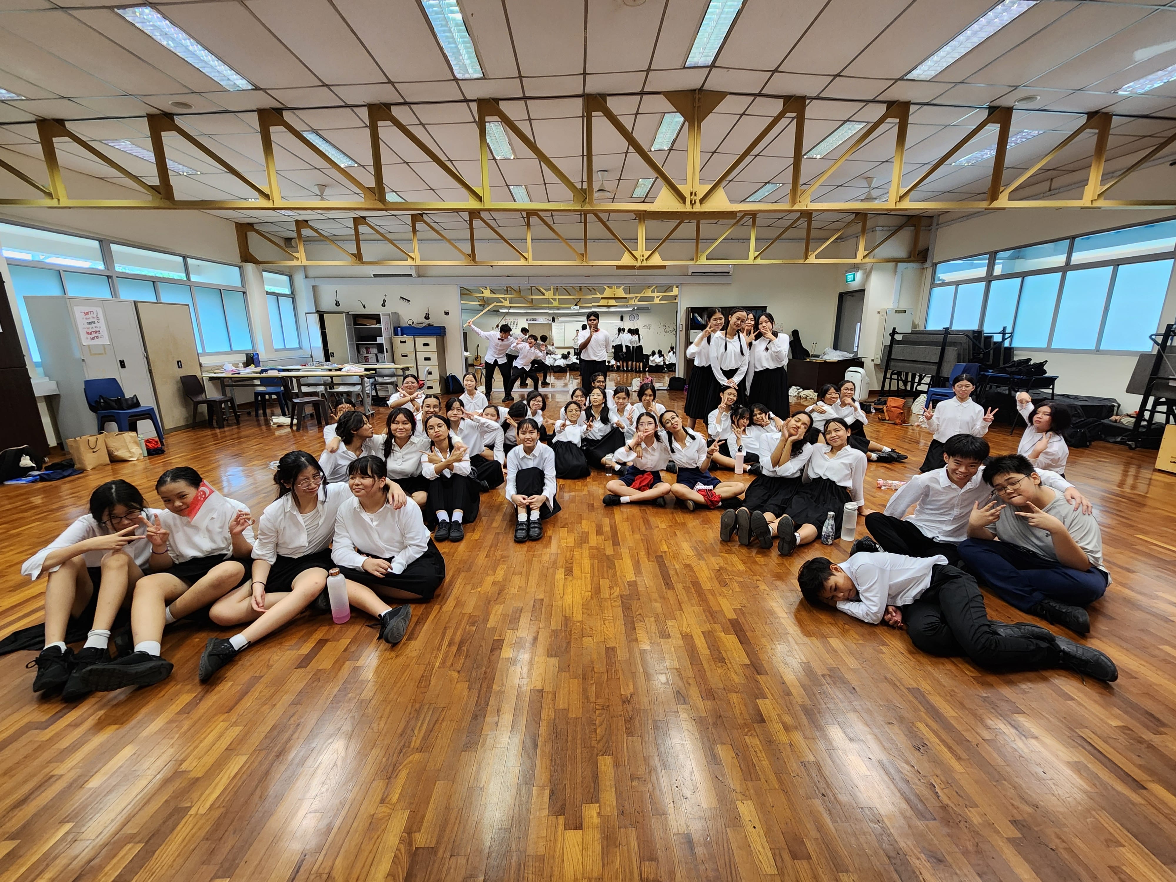 A large group of students in white shirts and black skirts sit and pose on a wooden floor in a school hall.