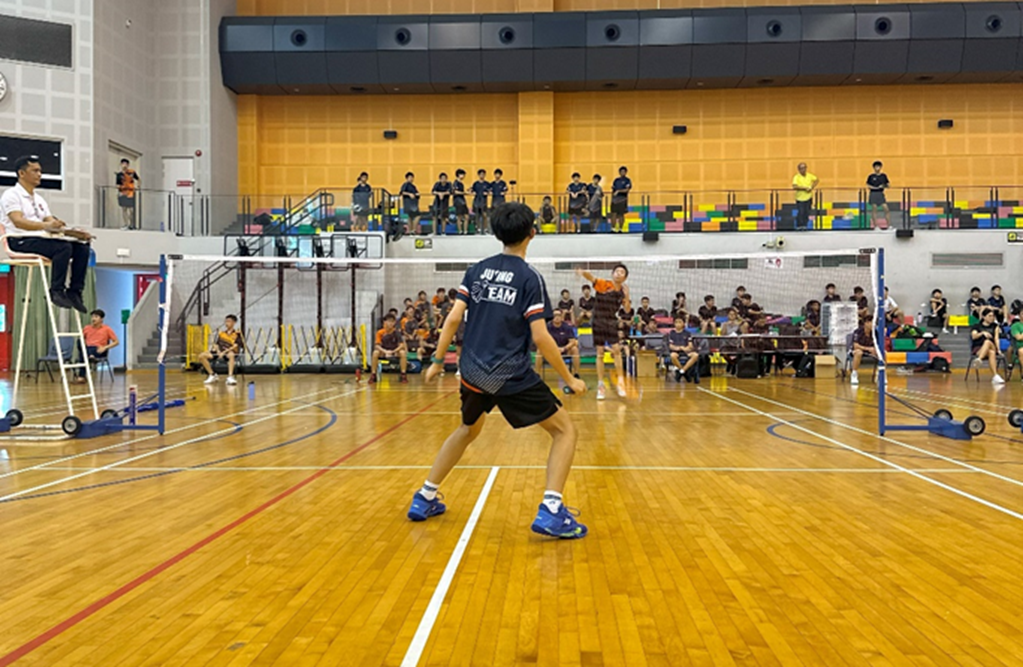 Two young players compete in a badminton match indoors with spectators watching from the stands.