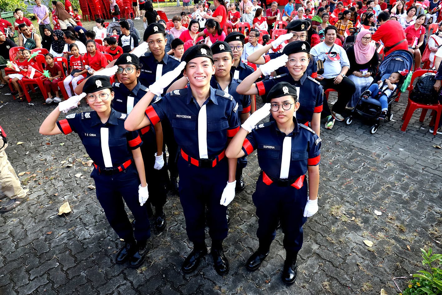 Six young cadets in navy and red uniforms salute outdoors, with a crowd in red seated behind them.