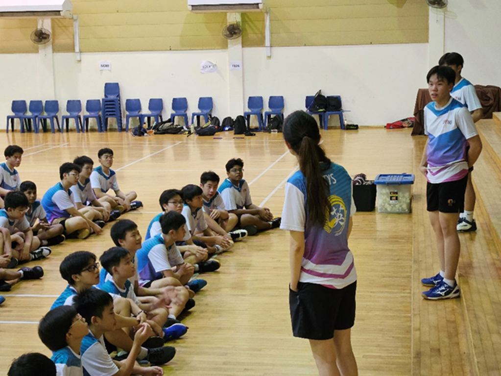 Two coaches in sports jerseys address a group of young students sitting on a gymnasium floor.