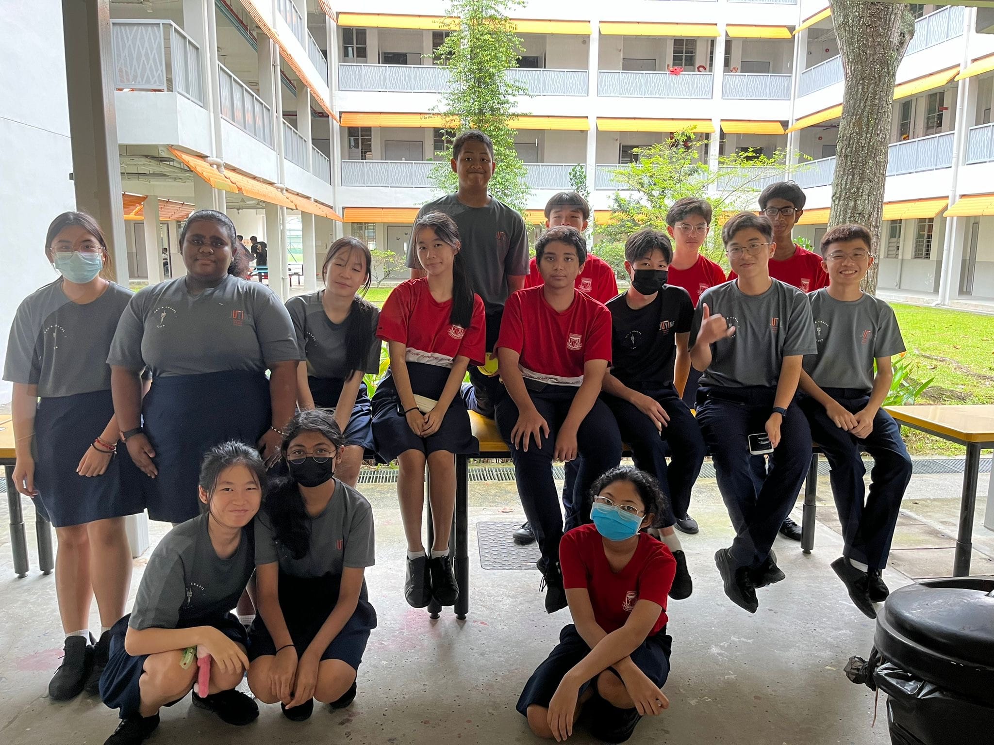 Group of teenagers in red and gray school uniforms posing together in an outdoor school courtyard.