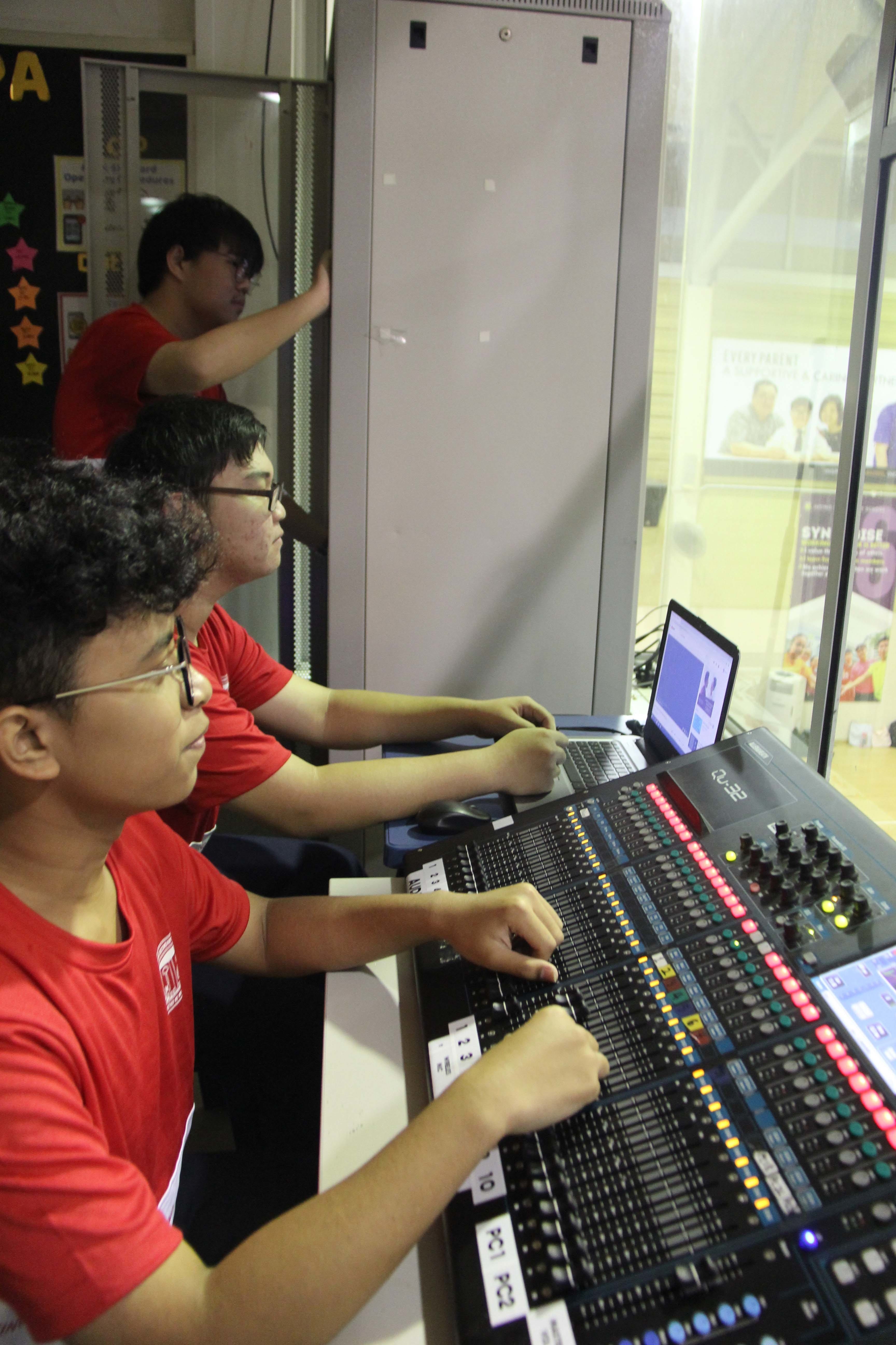 Three young people in red shirts operate a large audio mixing console with a laptop in a control room.