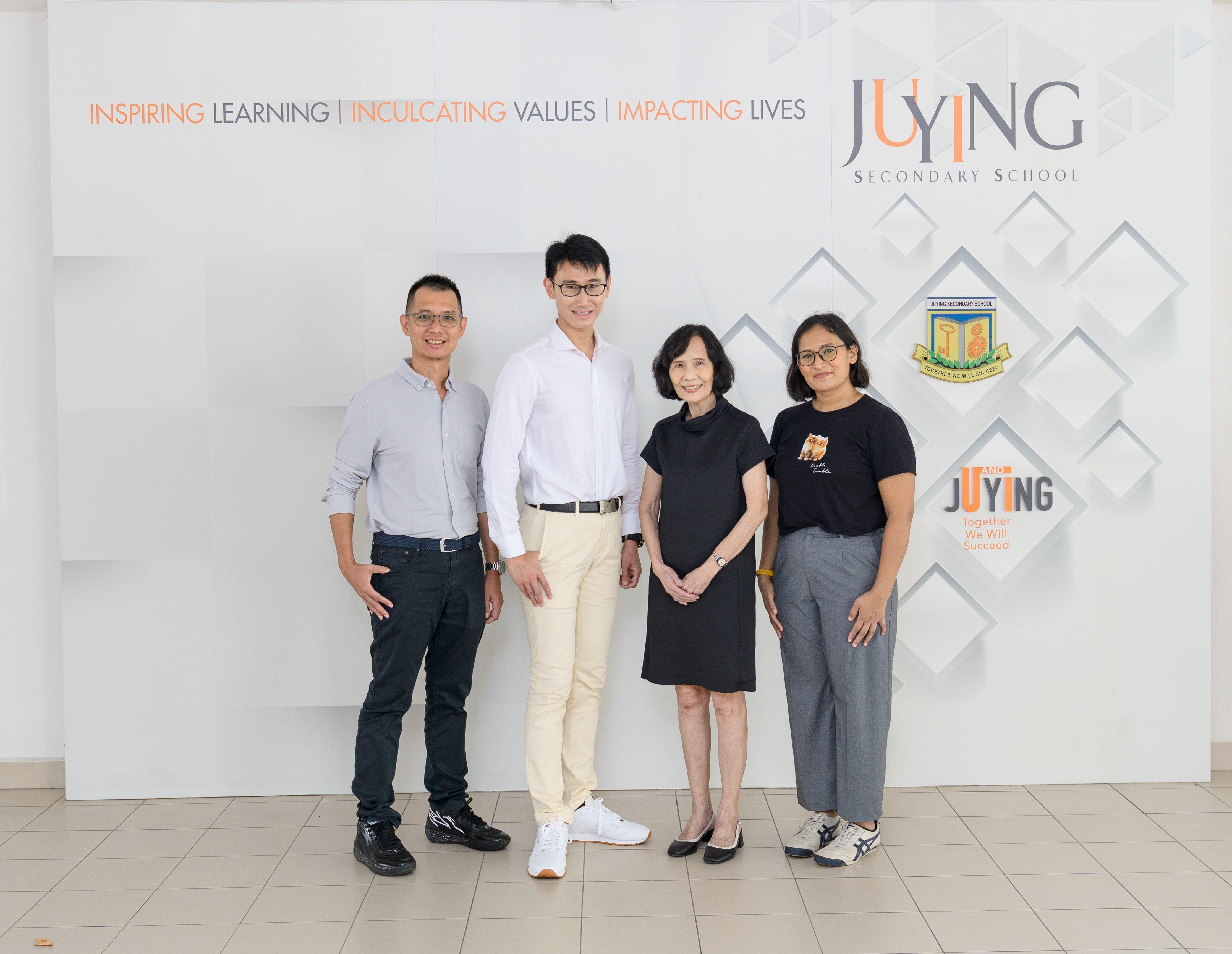 Four people posing in front of a Juying Secondary School banner reading Inspiring Learning, Inculcating Values, Impacting Lives.