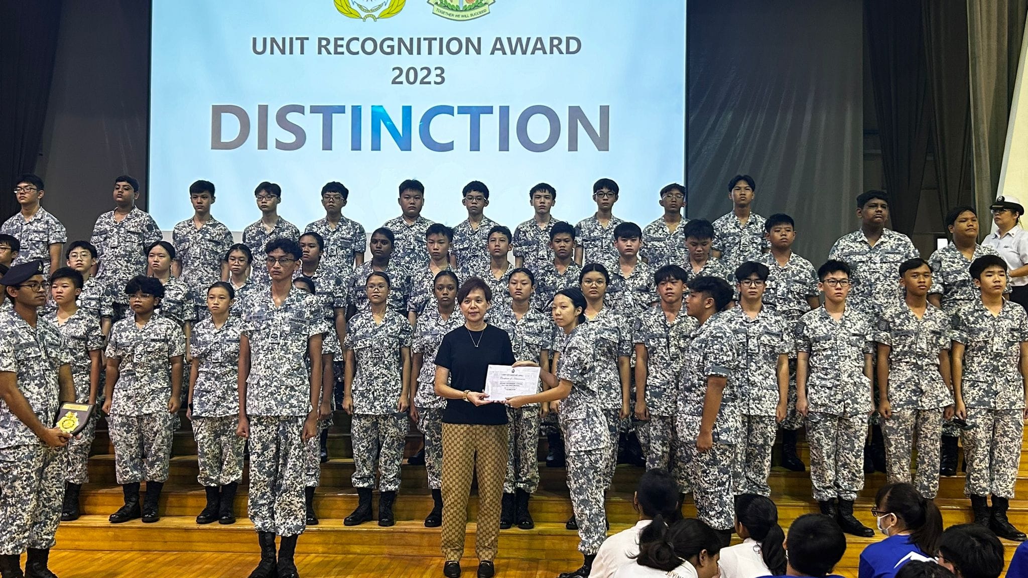 A group in camouflage uniforms stands on a stage behind a screen reading "Unit Recognition Award 2023 Distinction."
