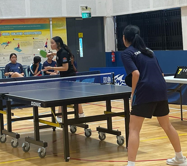 Two girls in navy uniforms play table tennis on a Donic table in an indoor sports hall.