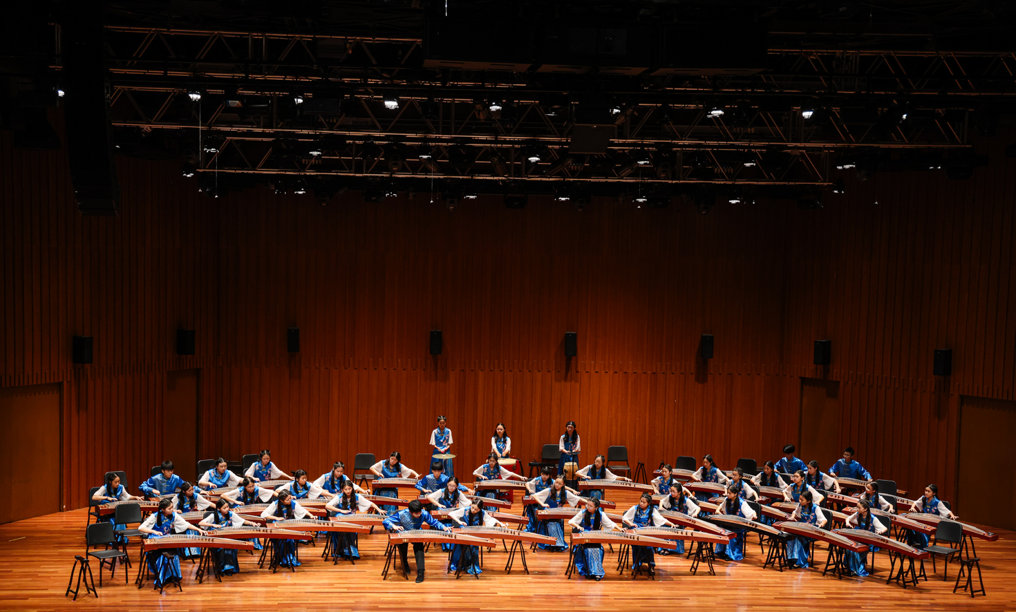 Large group of performers in blue and white attire playing guzheng instruments on a wooden concert stage.