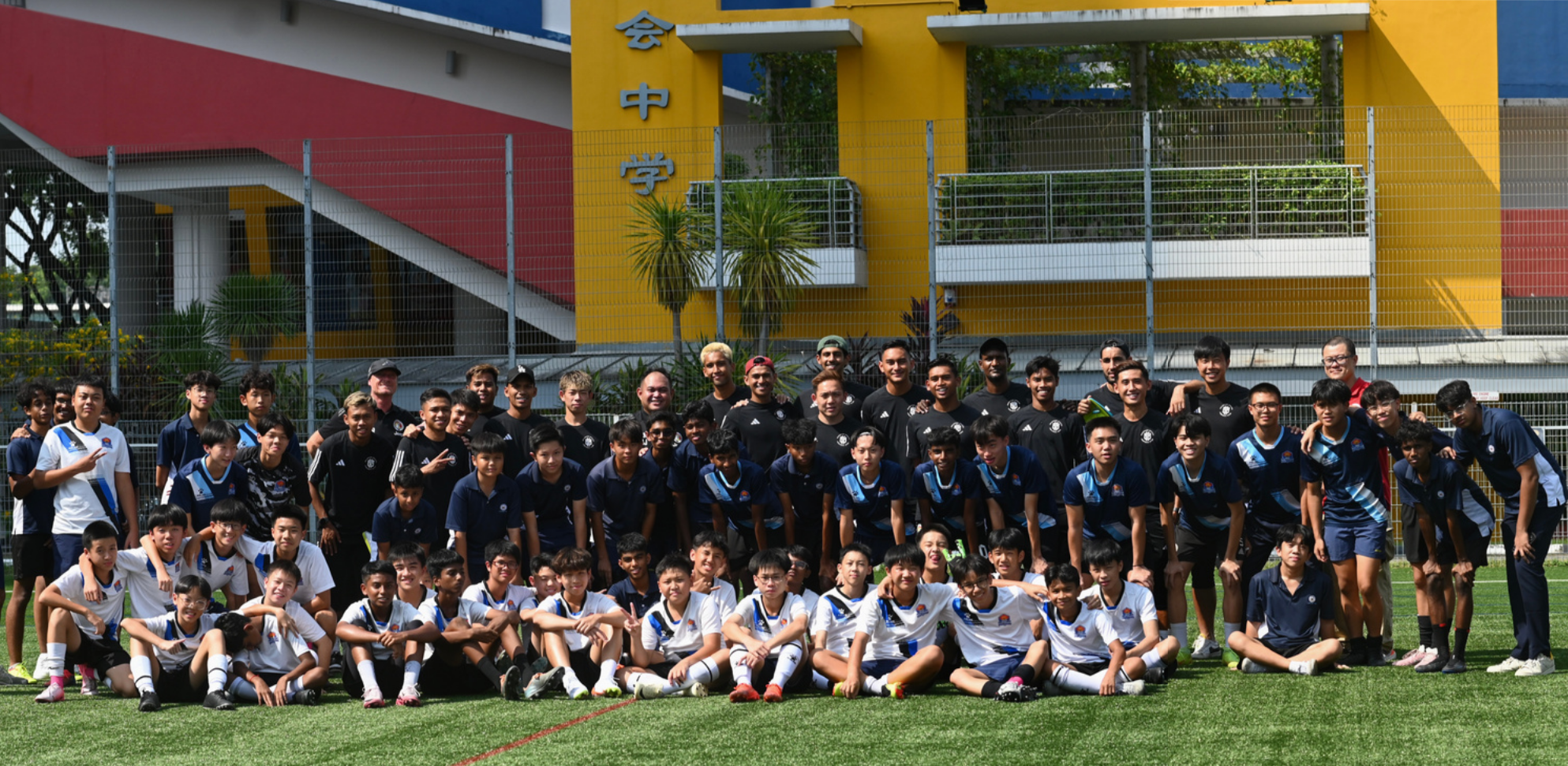 Large group of youth and adult football players posing on a green field in front of a colorful building with Chinese characters.