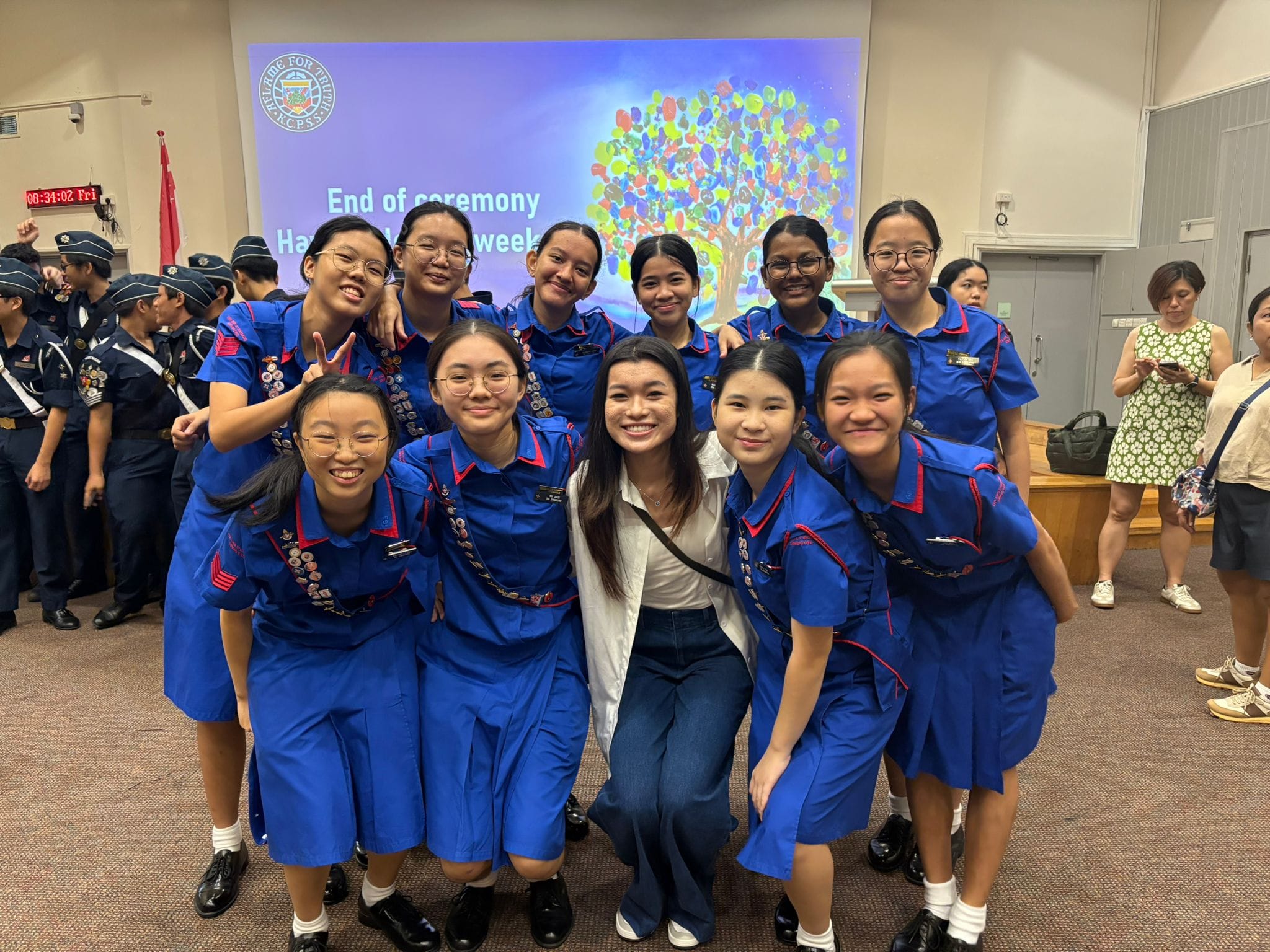 A group of young girls in blue uniforms pose smiling at an end of ceremony event with a colorful tree slide behind them.