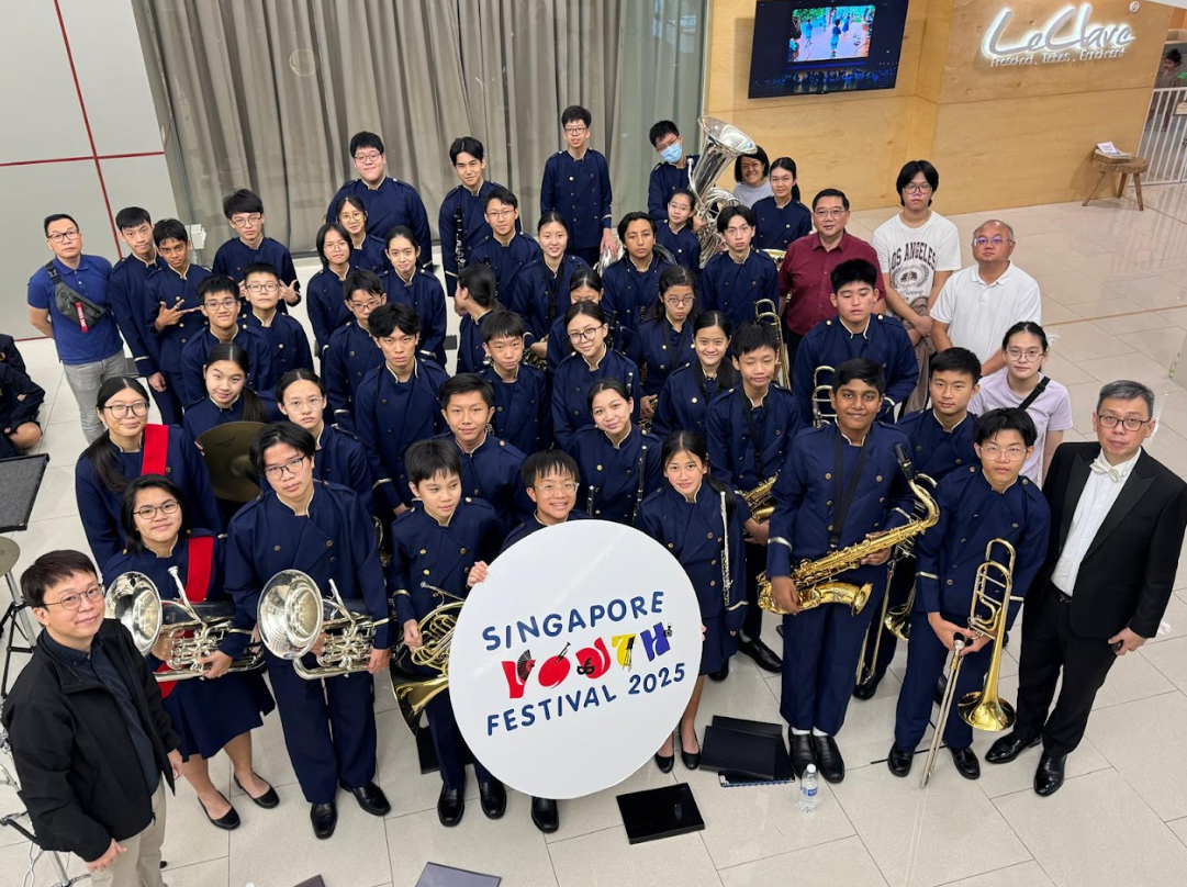 Youth band in navy uniforms holding brass instruments pose with a Singapore Youth Festival 2025 sign indoors.