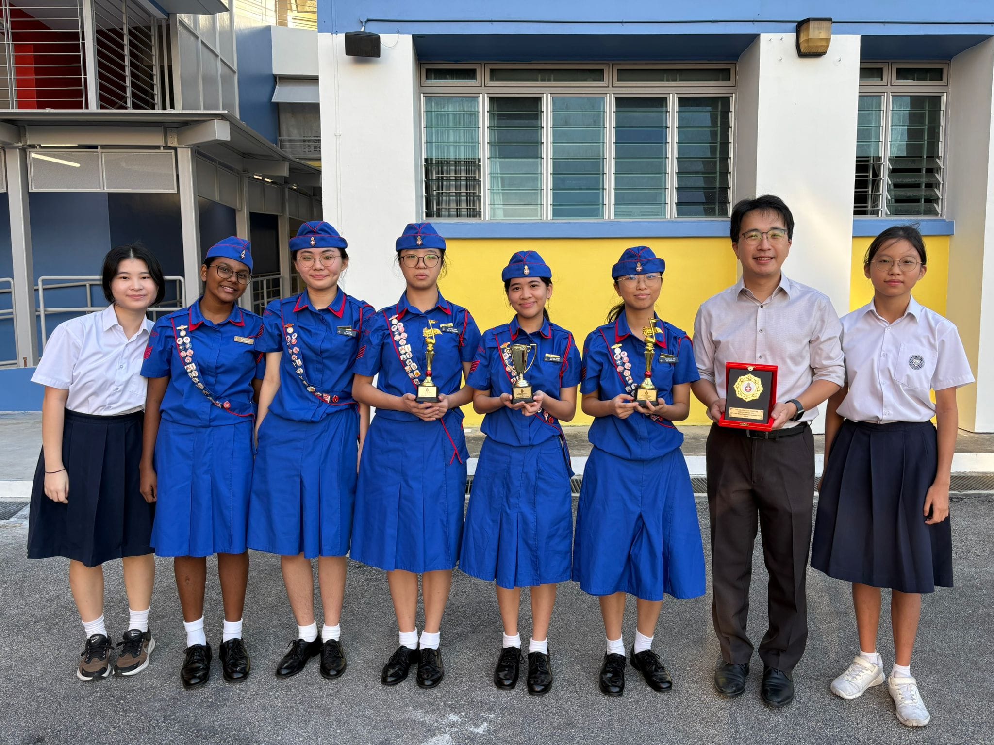 Seven people pose outdoors; four girls in blue scout uniforms hold trophies, flanked by two students and a man with a plaque.