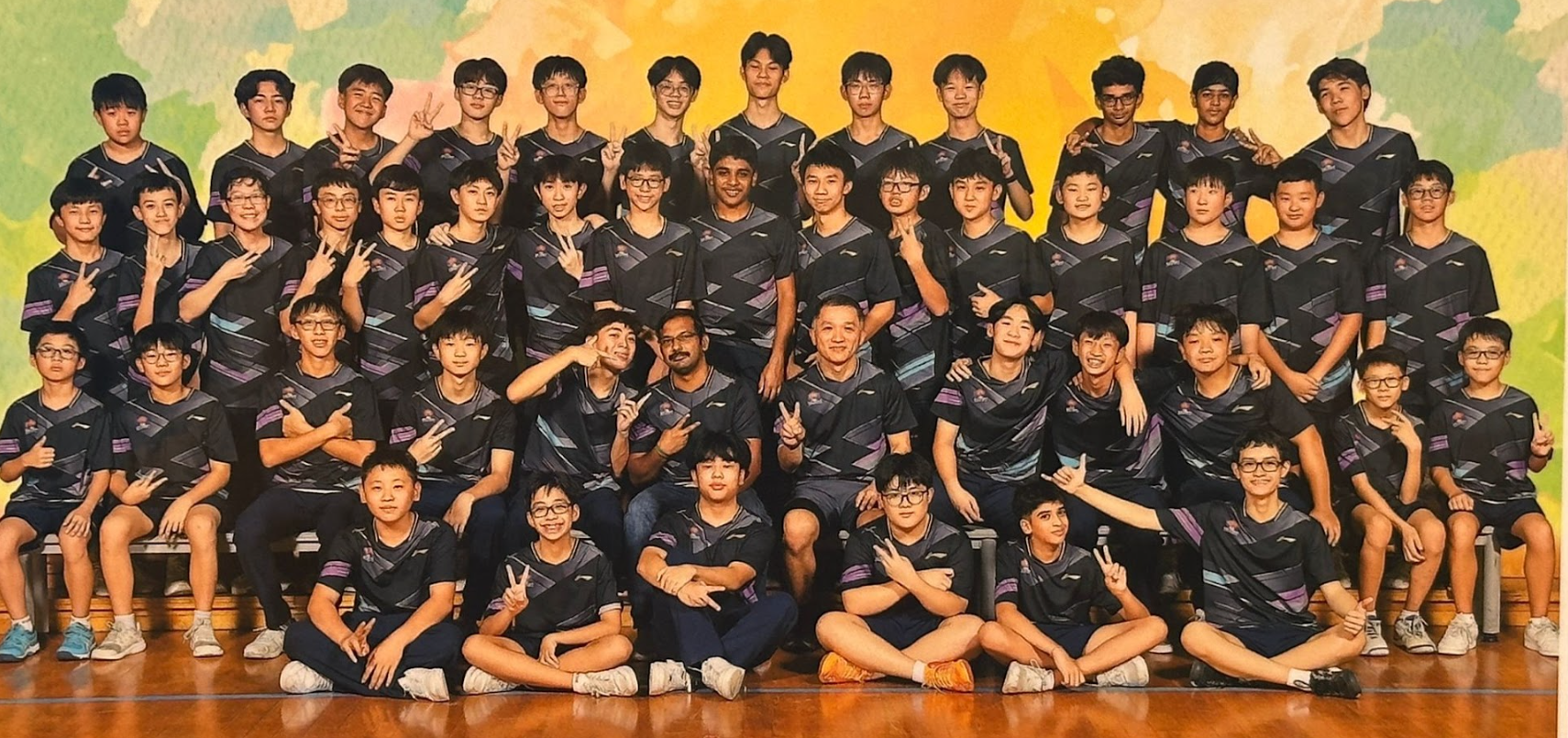 Large group of young male athletes in matching dark uniforms posing together in rows, many showing peace signs.