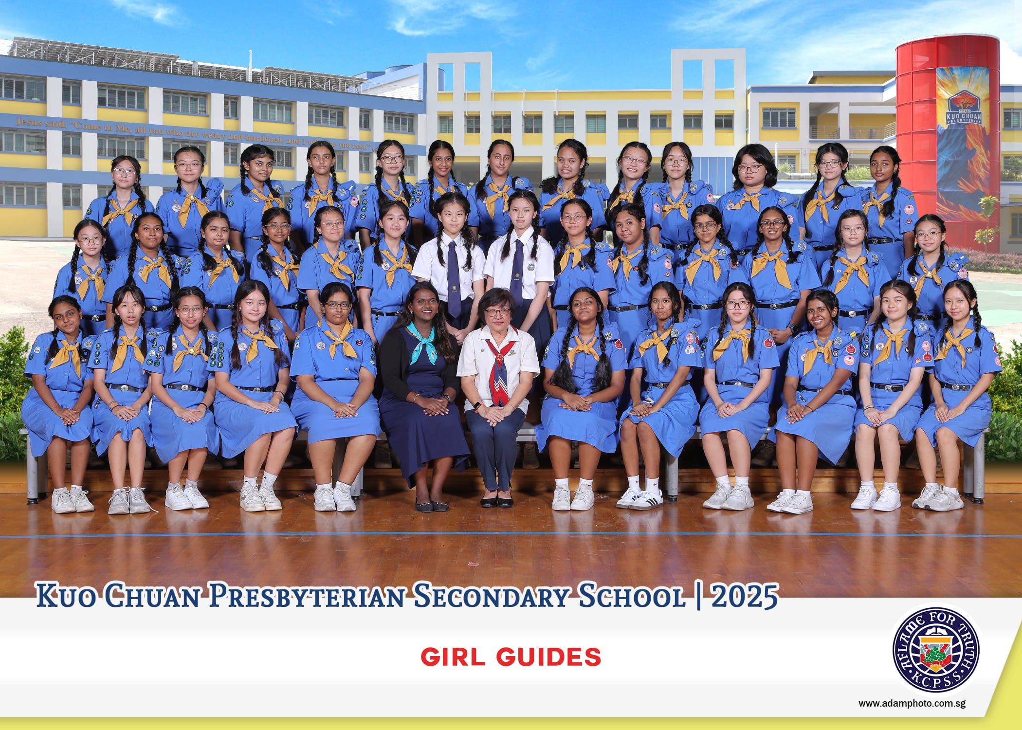 Group photo of Girl Guides in blue uniforms with leaders, posed before a school building, labeled 2025