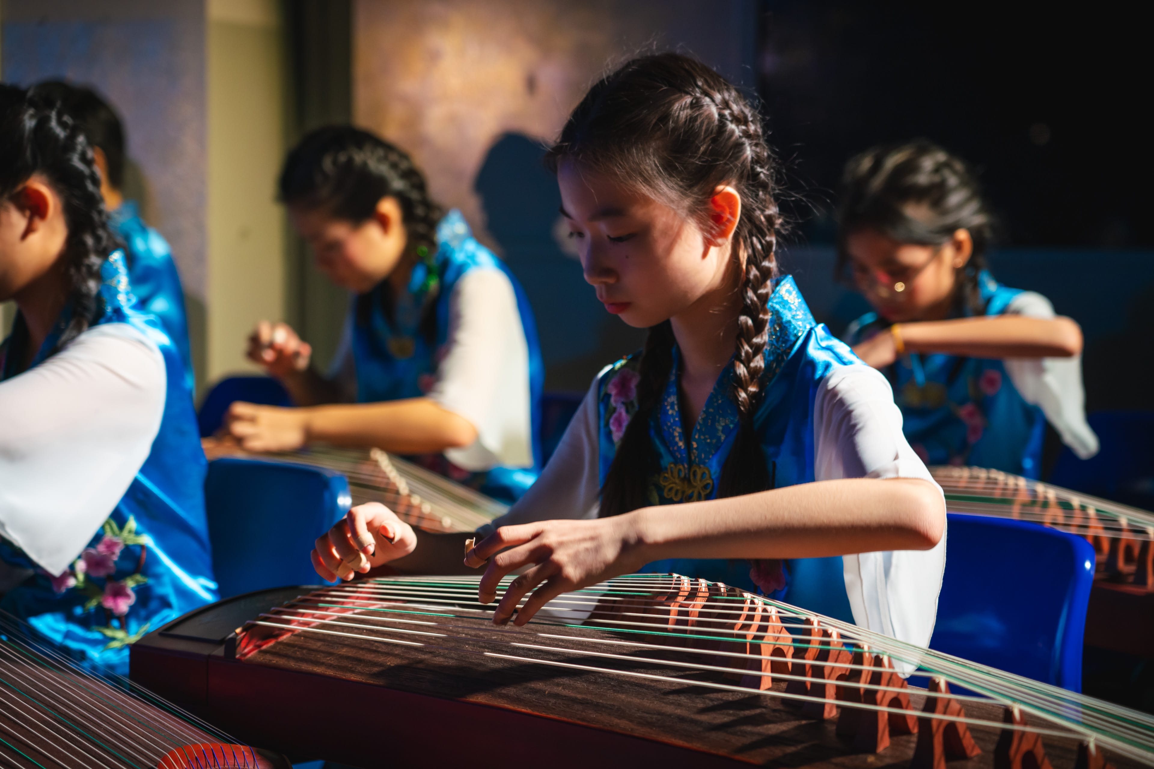 Young girls in blue embroidered outfits playing guzheng string instruments with focused expressions.