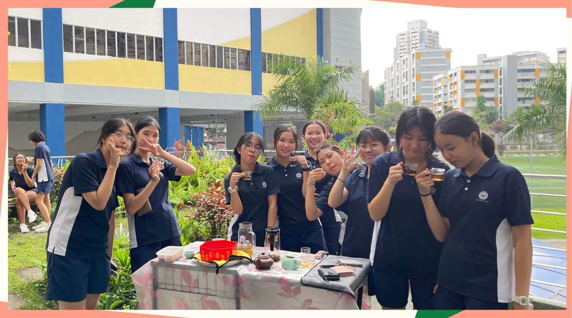 A group of students in navy uniforms pose outdoors holding small tea cups near a table with a tea set.