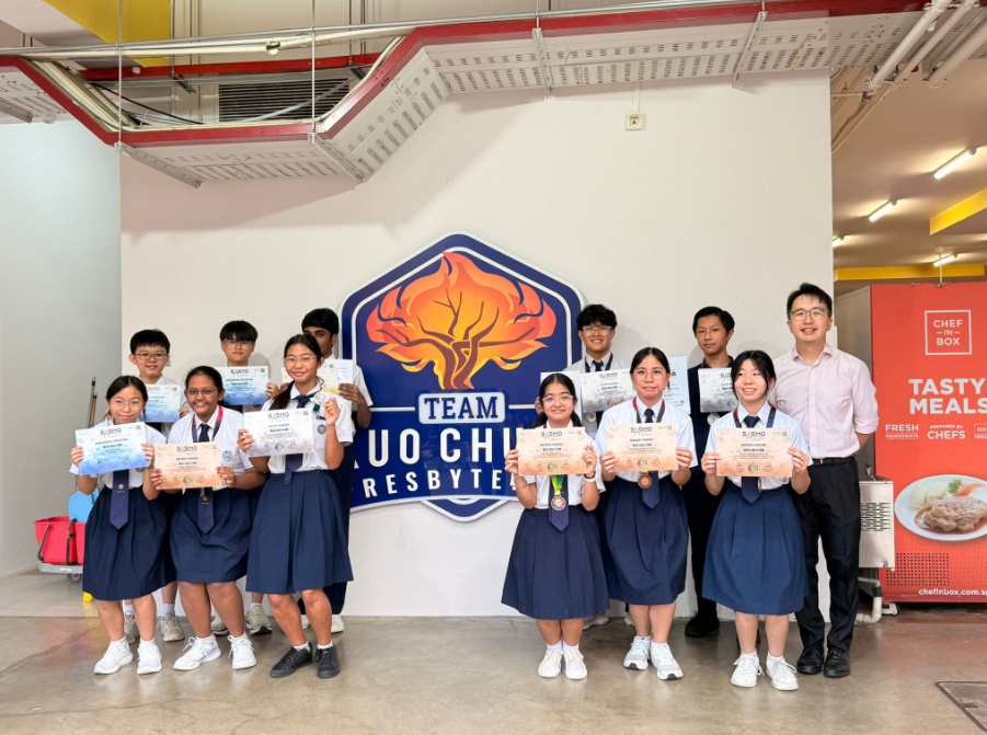A group of students in school uniforms holding certificates pose in front of a Team Kuo Chuan Presbyterian sign.