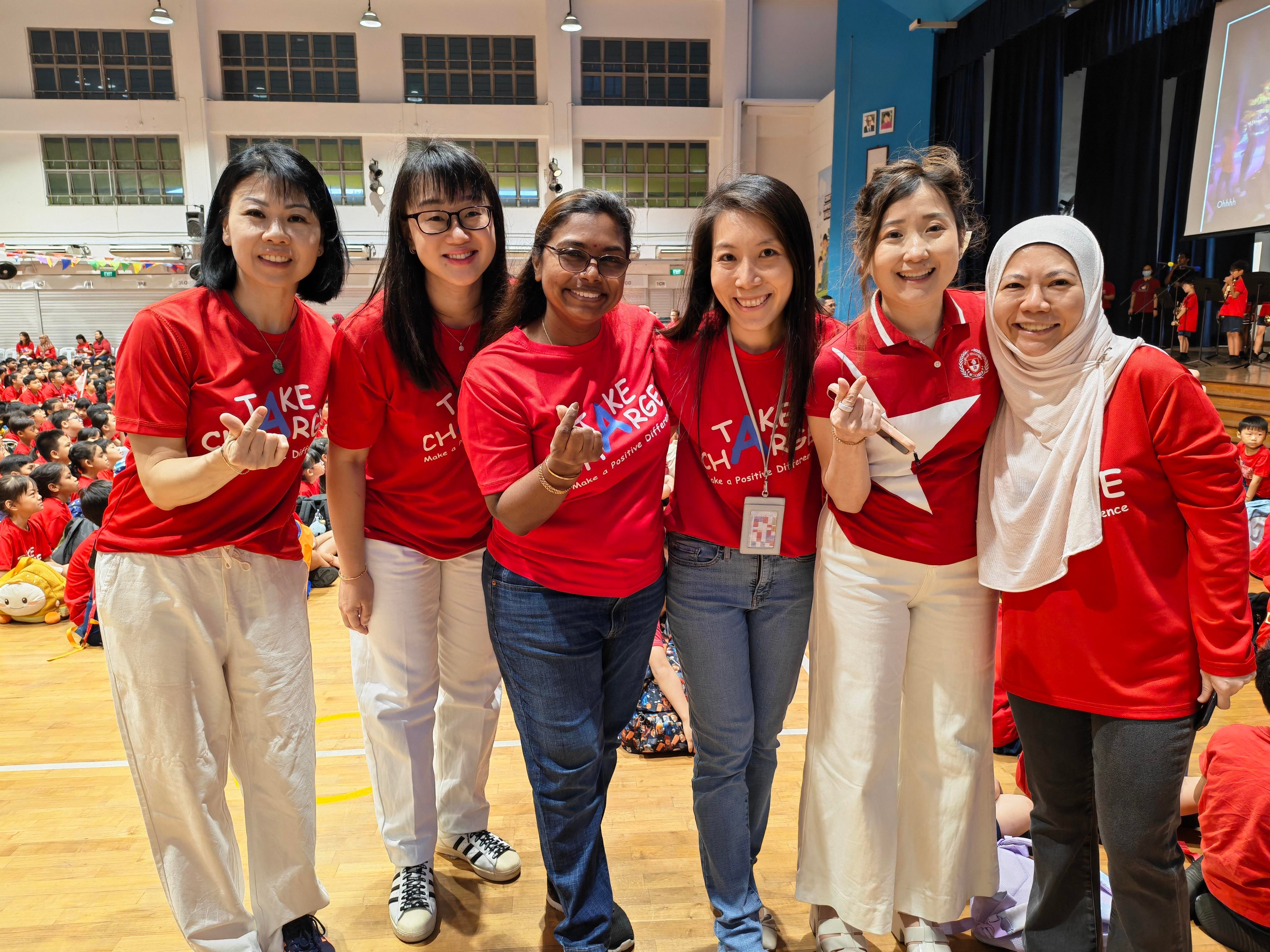 Six teachers dressed in red and white smile together in the school hall during the National Day celebrations, with students gathered for the concert in the background.