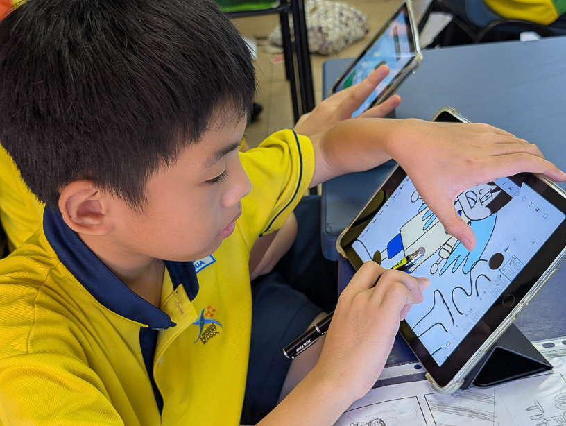 A young boy in a yellow school uniform draws on a tablet using a stylus in a classroom.