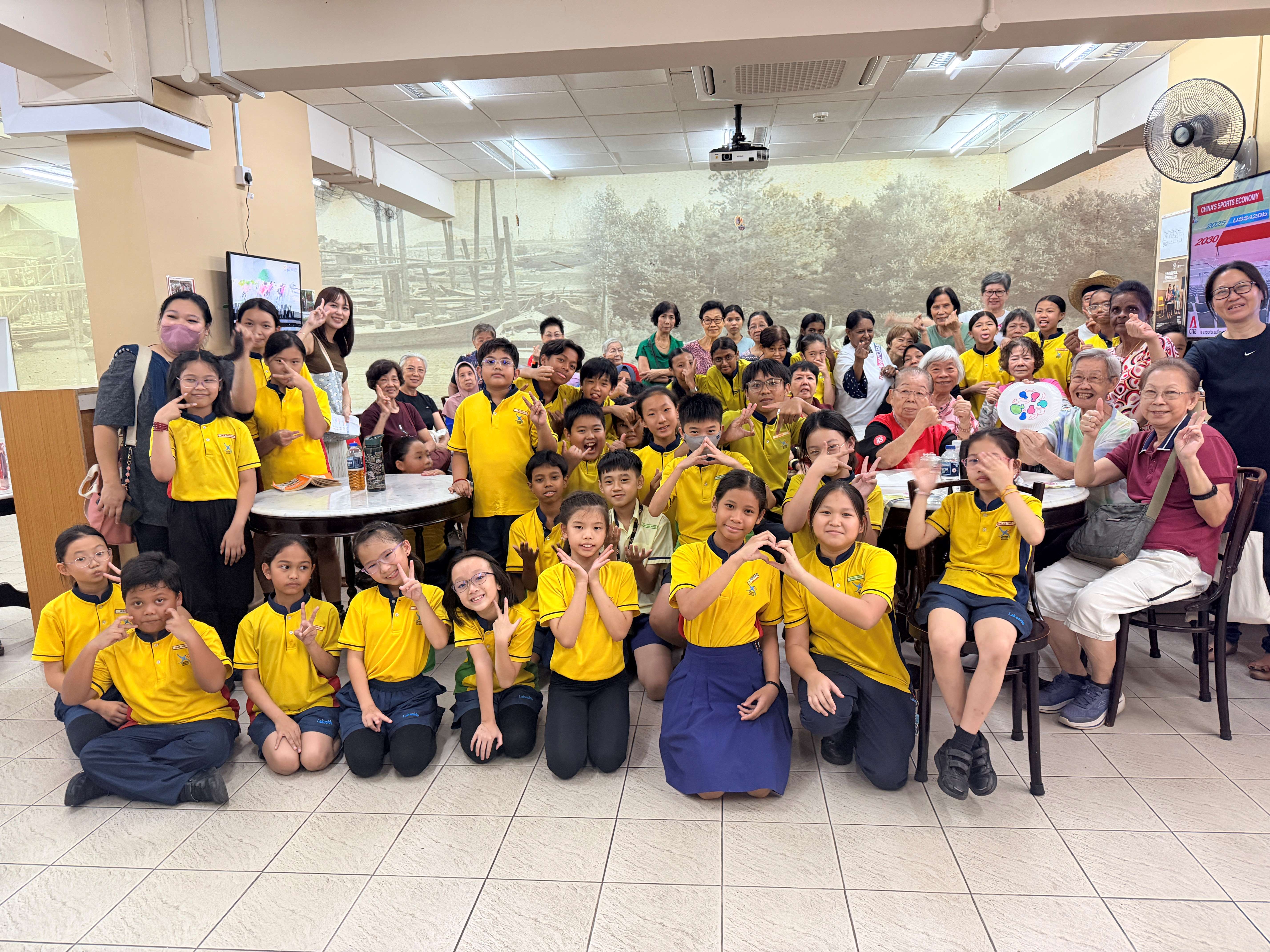 A large group of children in yellow uniforms and elderly adults pose together indoors, making hand gestures and smiling.