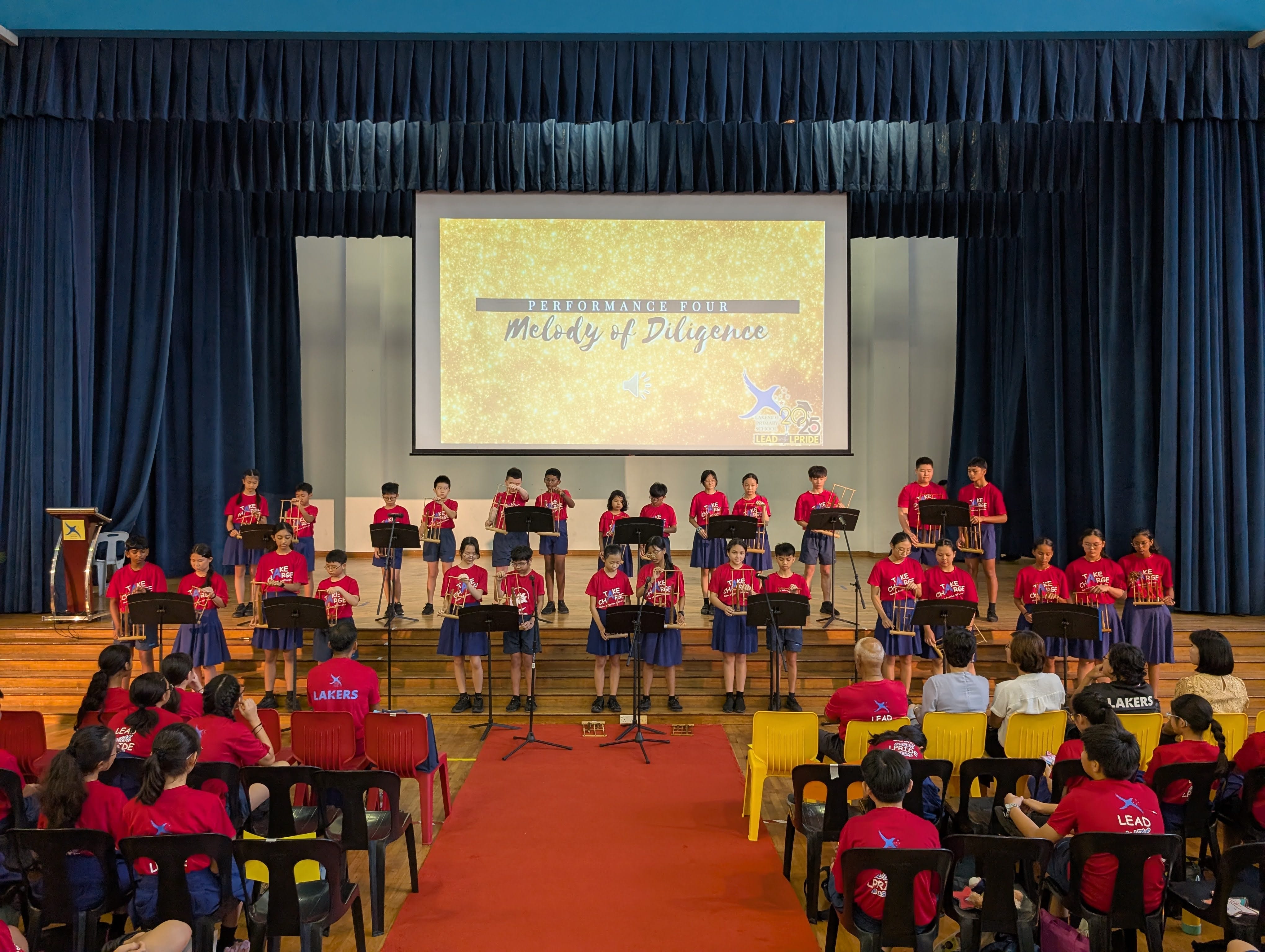 Students in red shirts perform on stage with music stands; a screen reads "Melody of Diligence" behind them.
