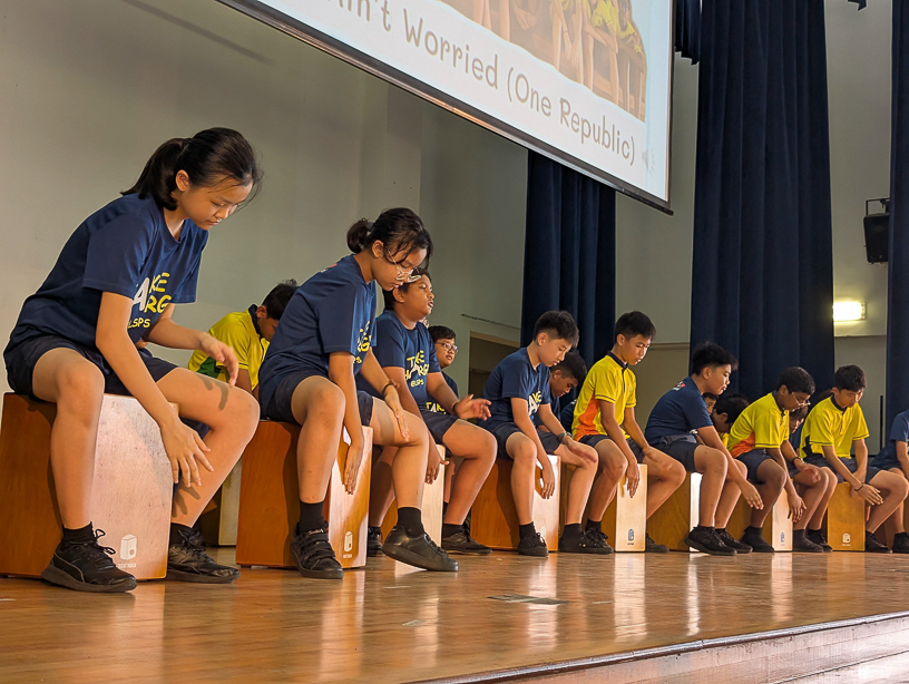Students in blue and yellow shirts sit on cajon drums on a school stage, performing percussion music.