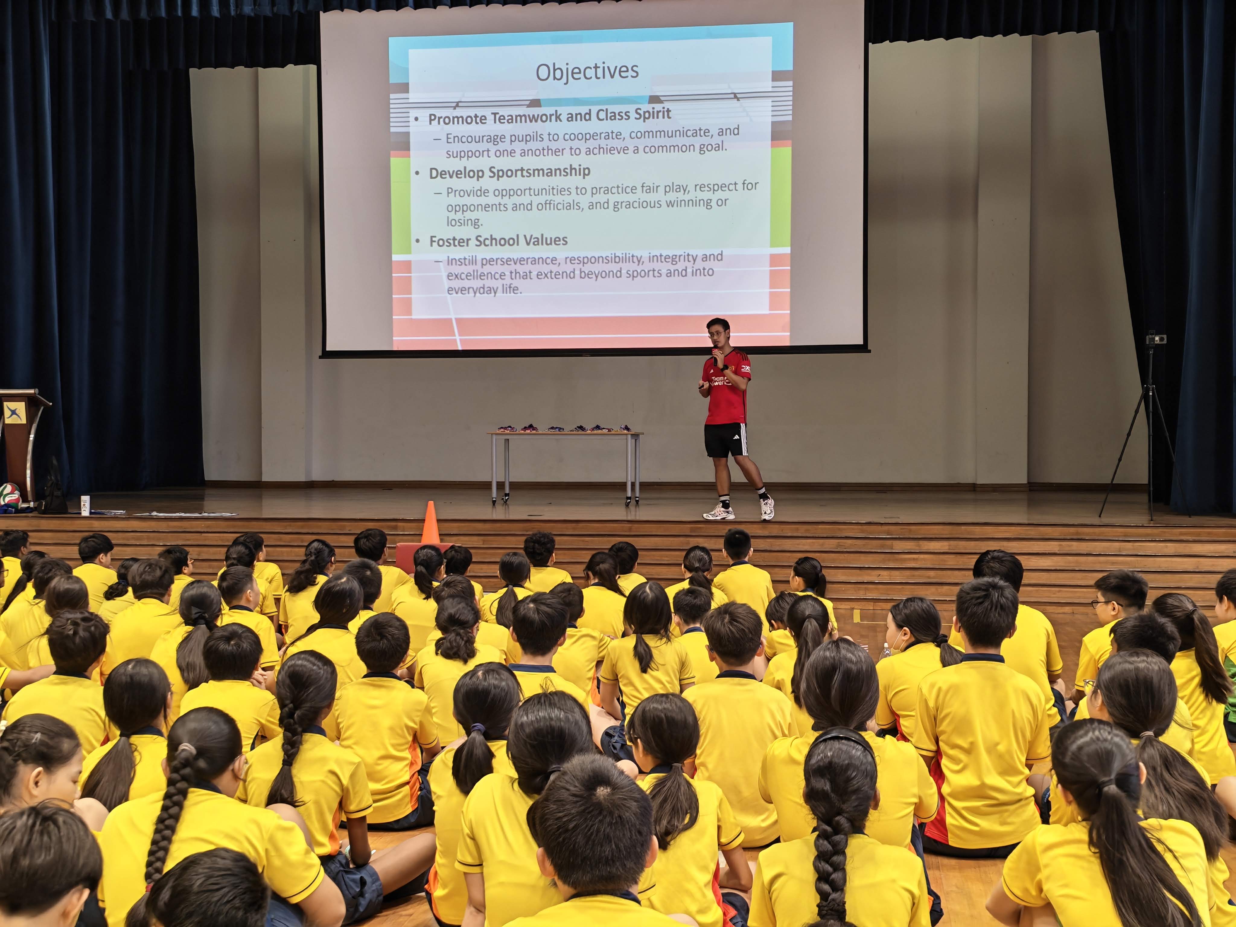All the students are gathered and seated as a teacher talks about displaying school values and sportsmanship during the games