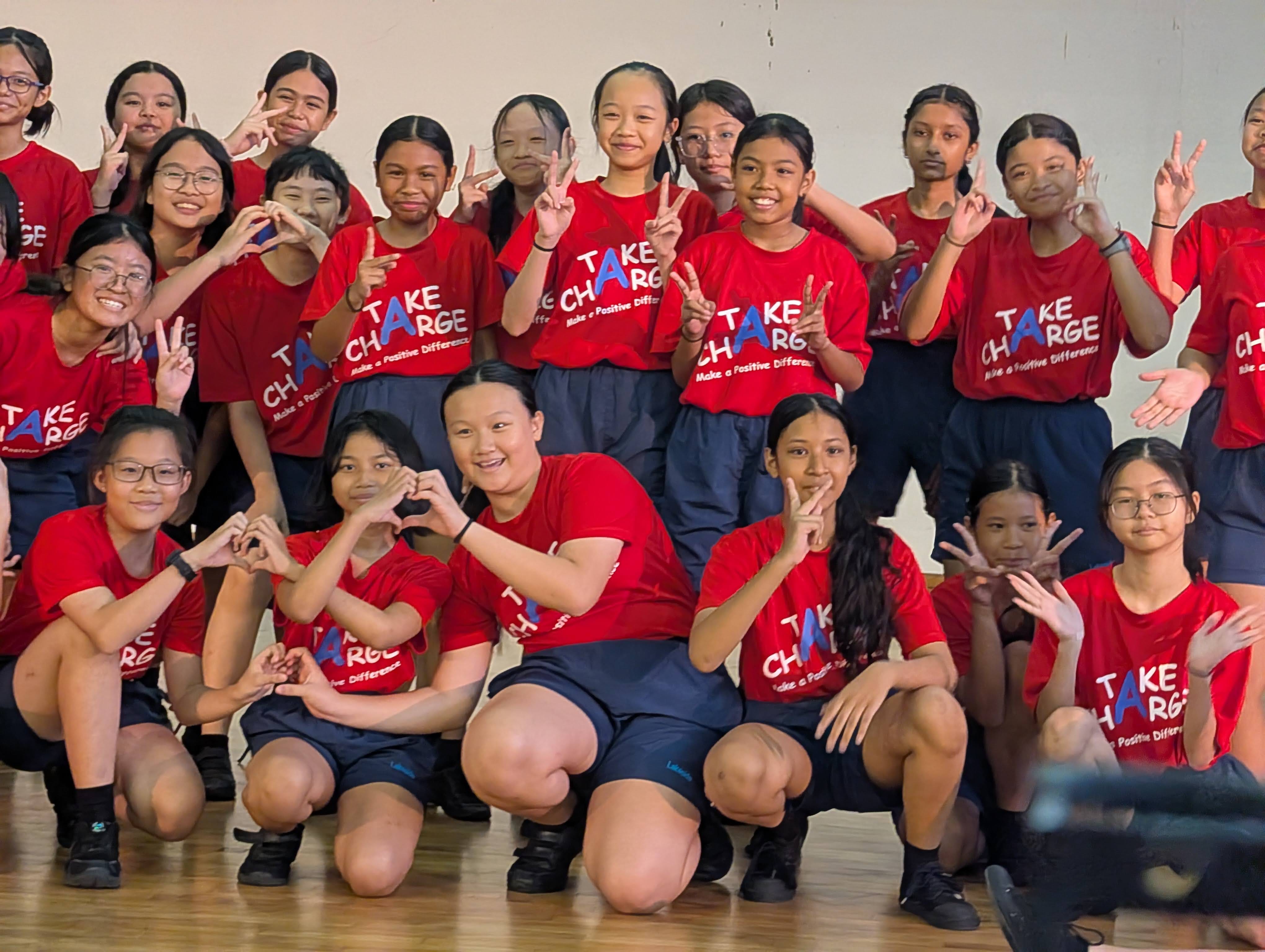A large group of girls wearing red Take Charge shirts pose with peace signs and heart hand gestures indoors.