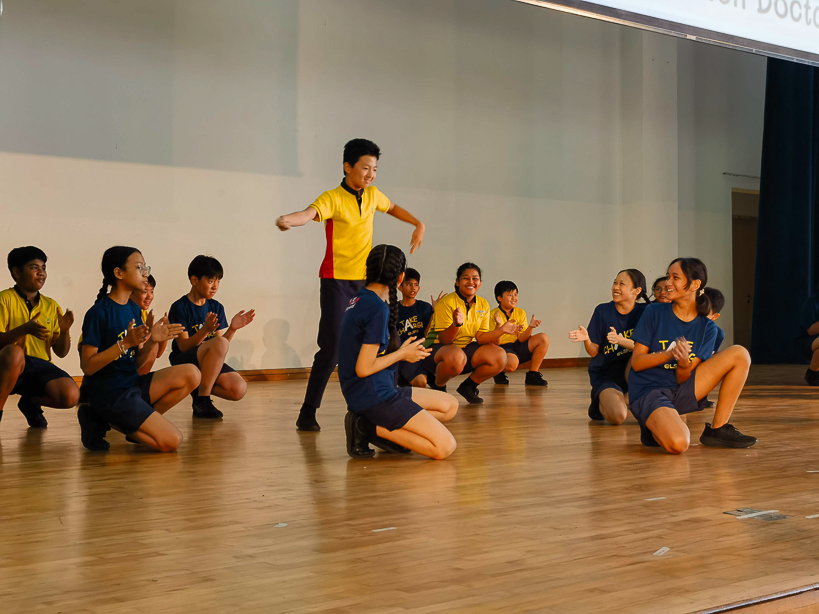 A boy in a yellow shirt dances on stage while classmates in blue and yellow uniforms clap and cheer around him.