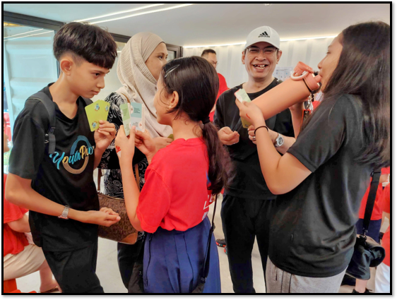A group of children and adults gathered indoors, holding and exchanging small cards while smiling.