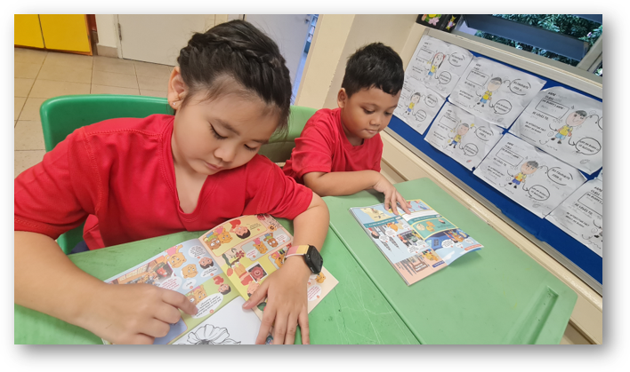 Two children in red shirts sit at a green table reading a colorful comic book: Kindsville Times Adventure  in a classroom.