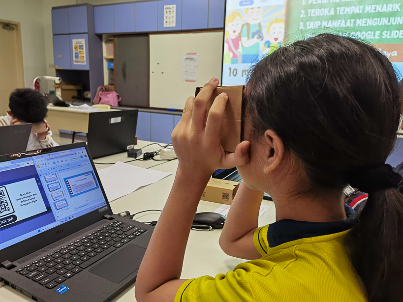 A girl in a yellow shirt holds a cardboard VR viewer to her face while sitting at a desk with a laptop in a classroom.