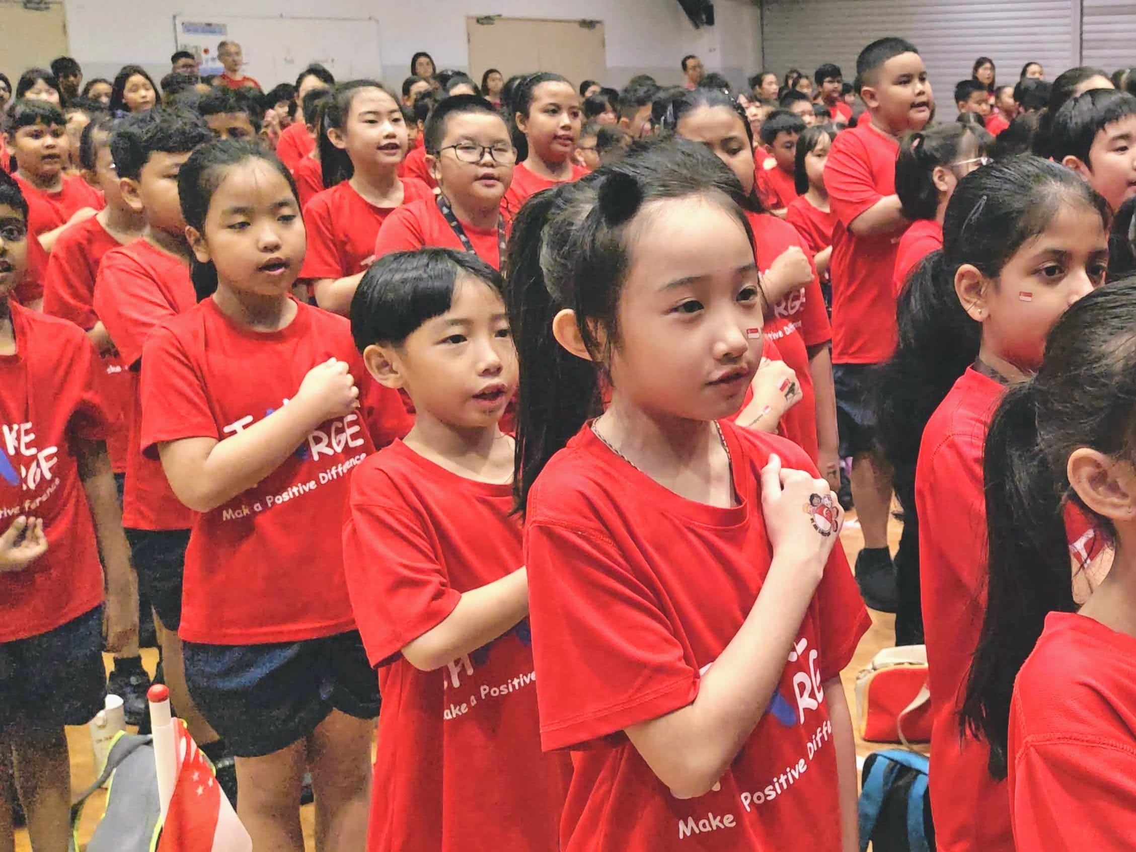 Students dressed in red stand together in the school hall with their right fists placed over their hearts as they recite the National Pledge during the National Day Observance Ceremony.