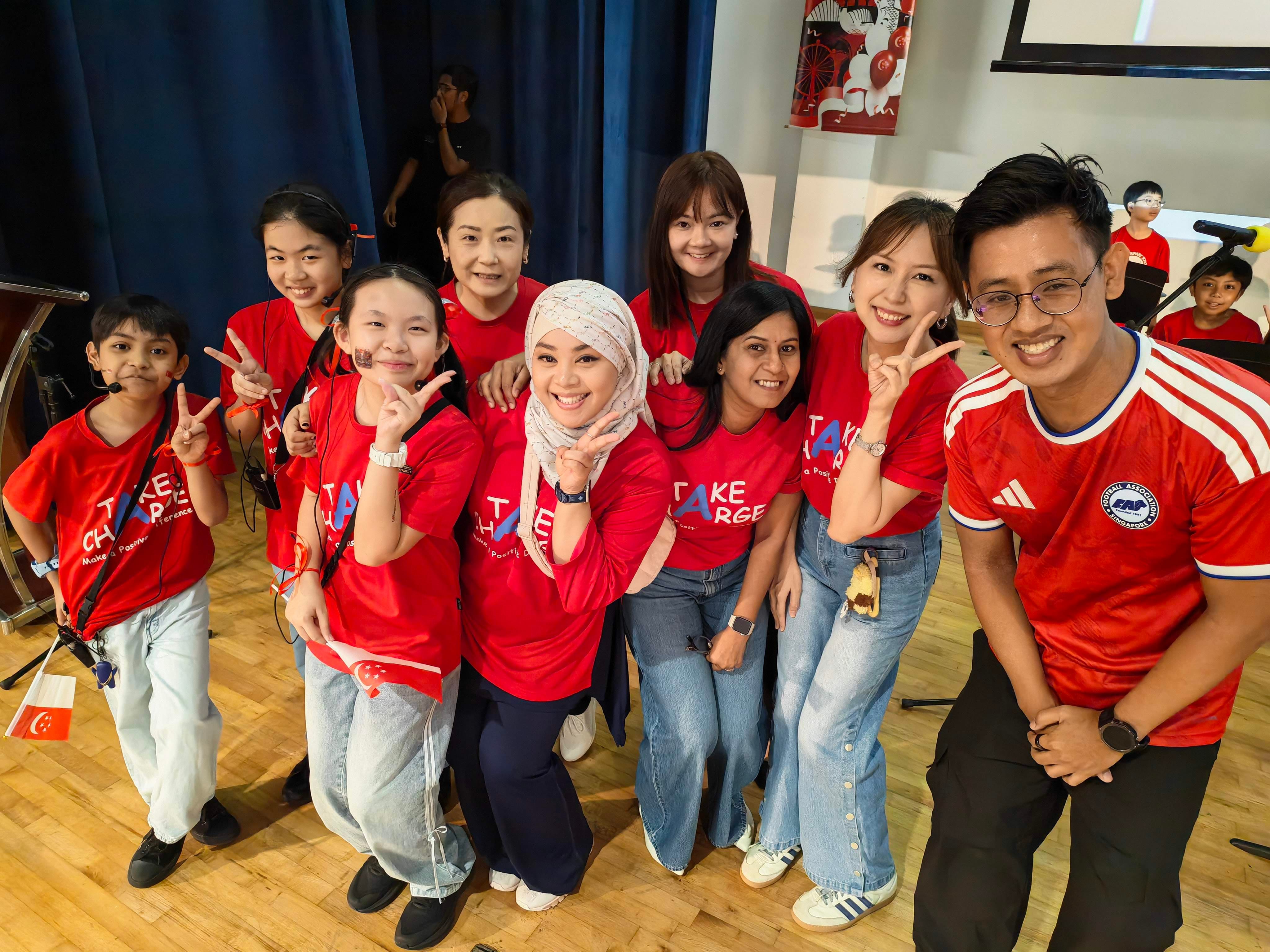 A group of teachers and student emcees dressed in red pose together on stage after working together to organise and host the school’s National Day concert.
