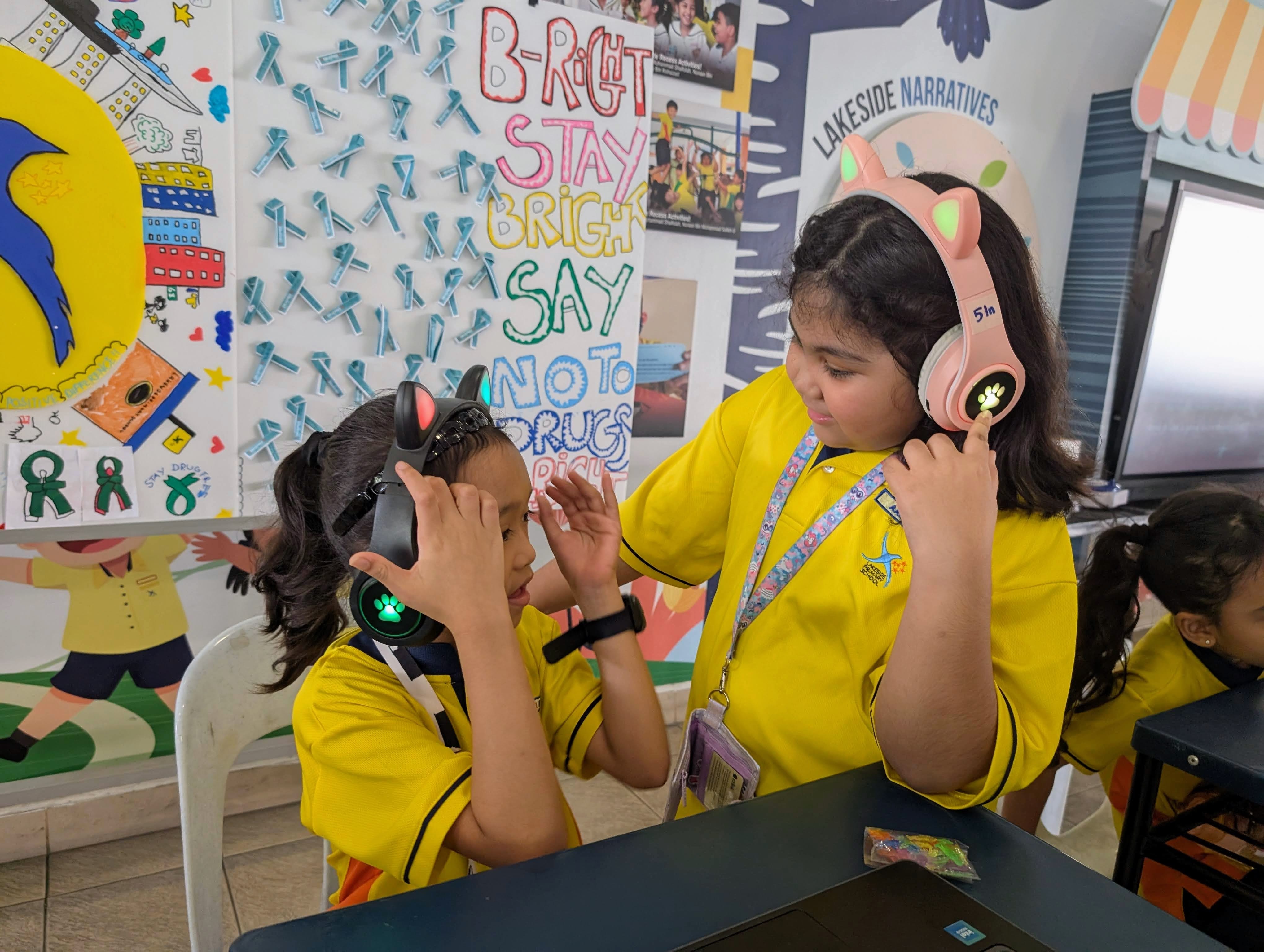 Two schoolgirls in yellow uniforms wear cat-ear headphones near a colorful "Say No to Drugs" poster.