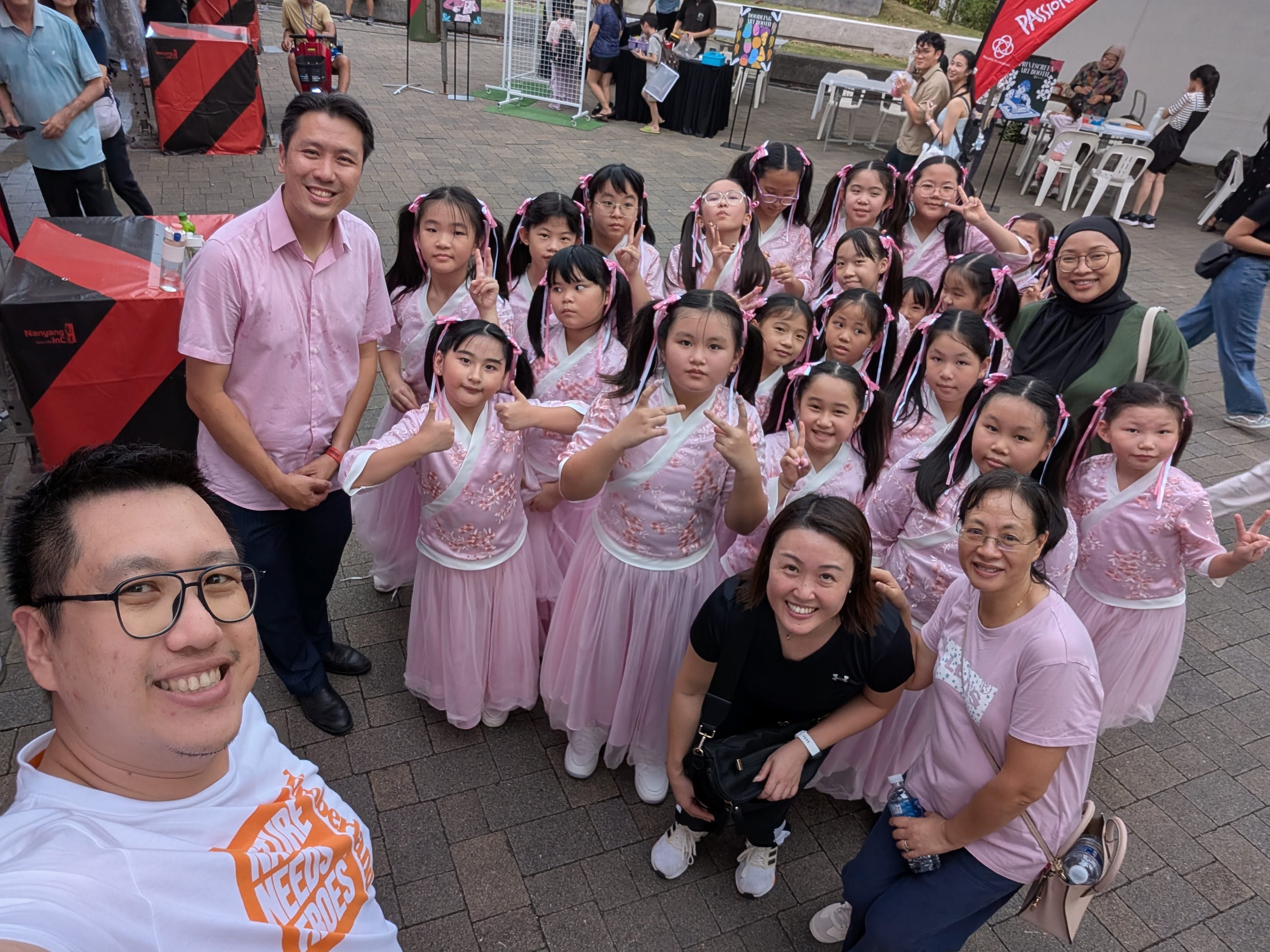 A group of young girls in pink traditional Chinese dresses pose with adults at an outdoor community event.
