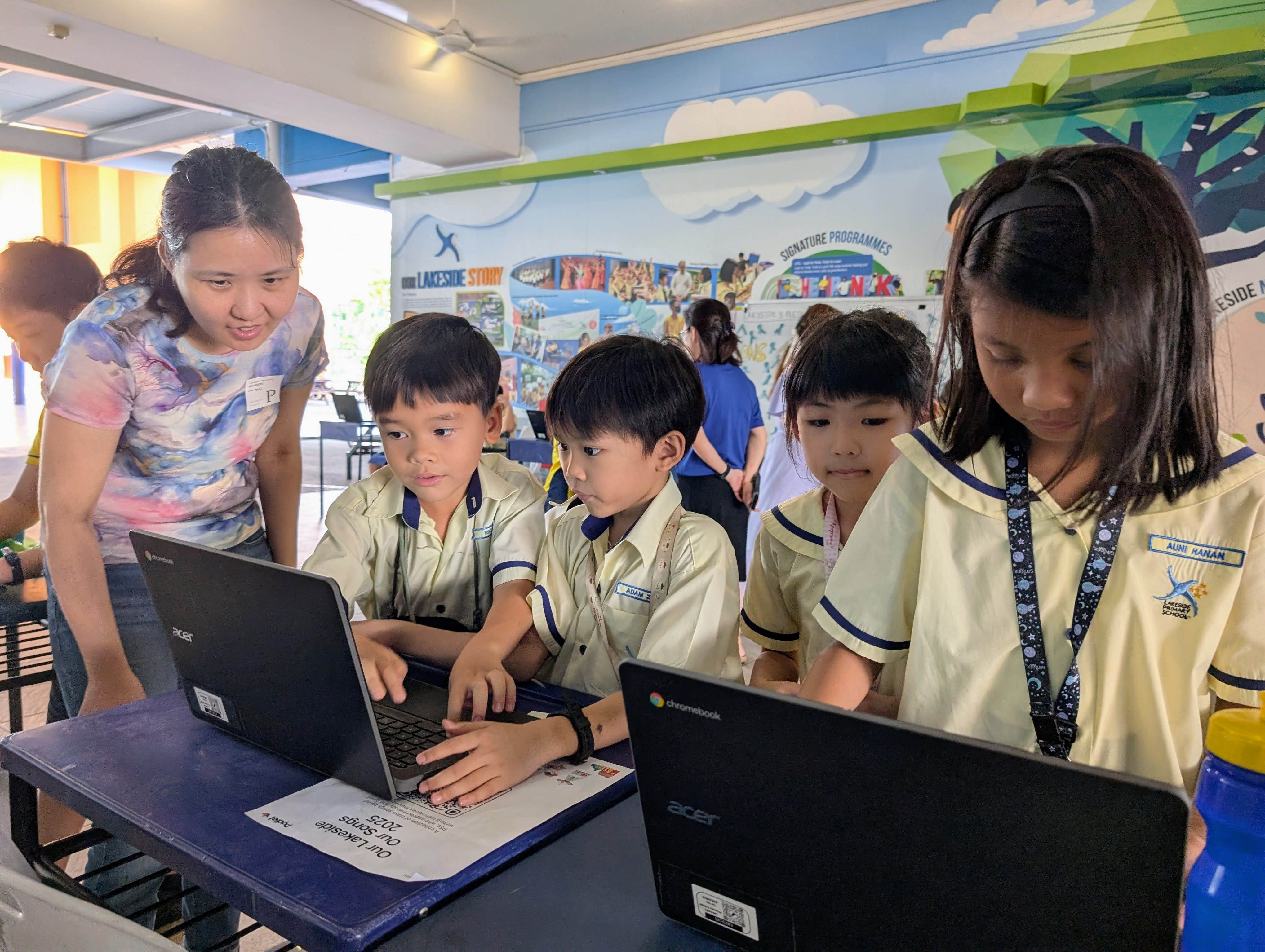Four young students in uniforms work on laptops while an adult woman leans over to guide them at school.