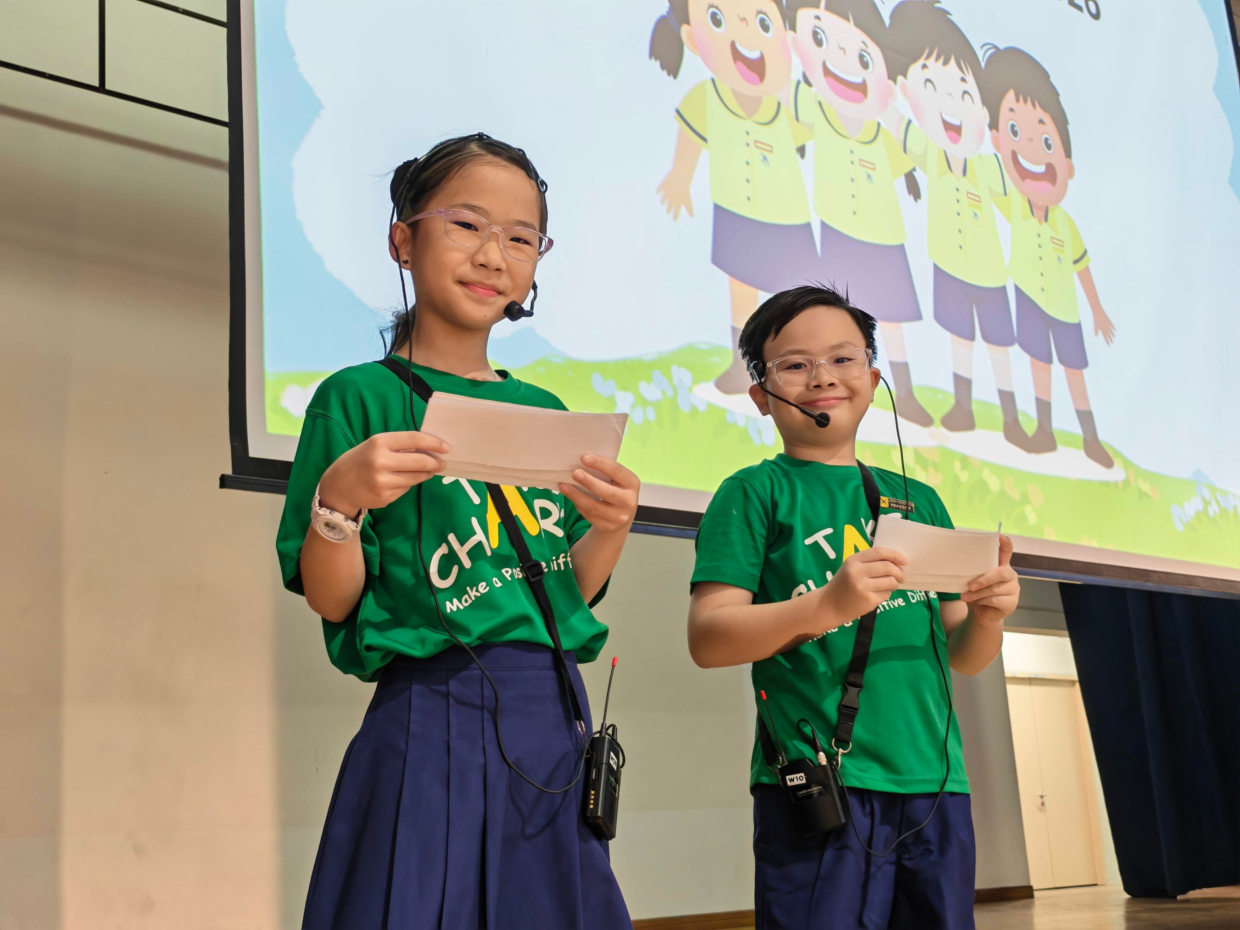 Two student presenters host the Racial Harmony Day assembly on stage, speaking into microphones while introducing the programme.