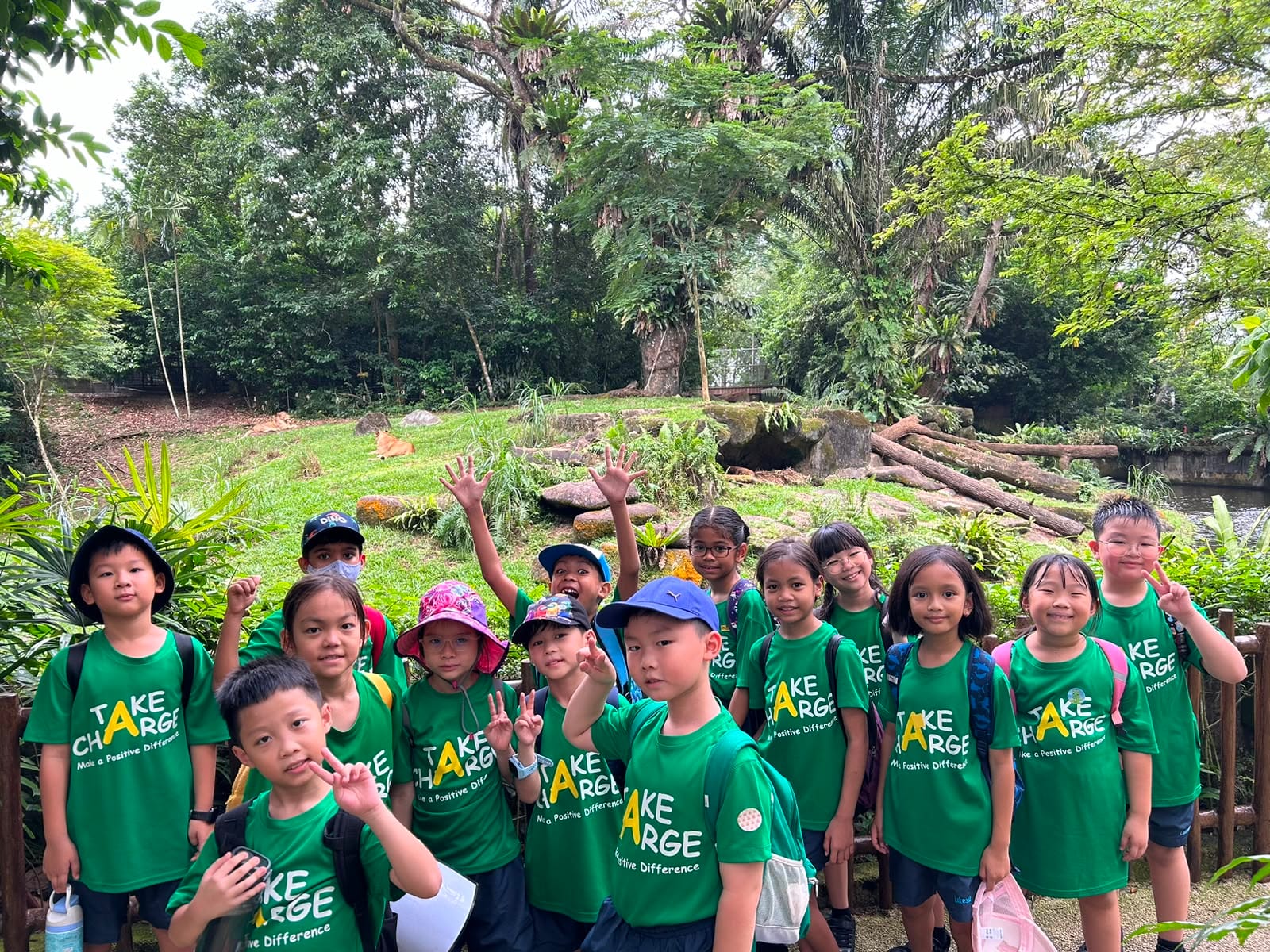 A group of students in green T-shirts posing and waving in front of a lion enclosure surrounded by lush greenery.