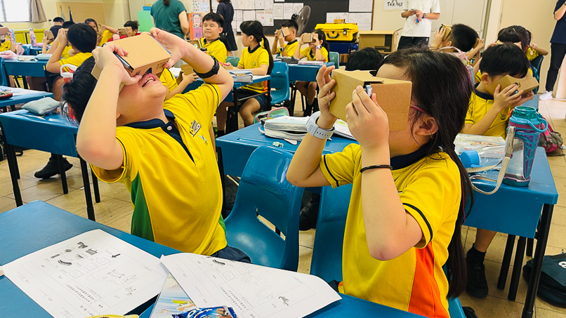 Students in yellow uniforms sit at blue desks using cardboard VR viewers held up to their faces in a classroom.